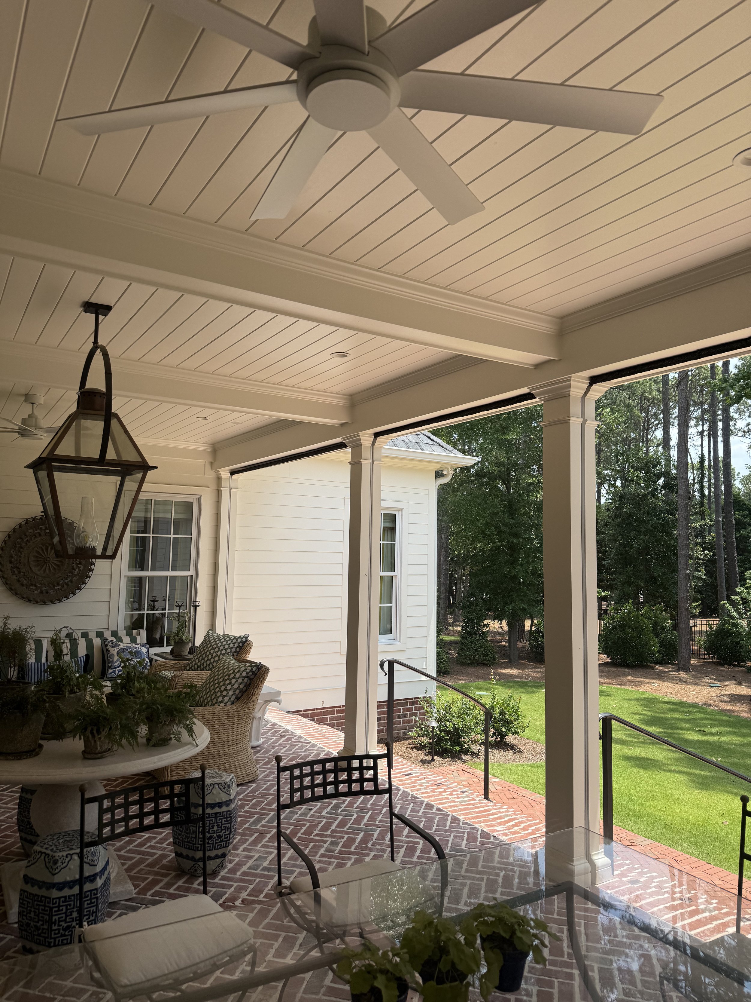 A covered porch with a ceiling fan, hanging lantern, and patio furniture including a glass table and chairs, overlooking a yard with grass and trees.