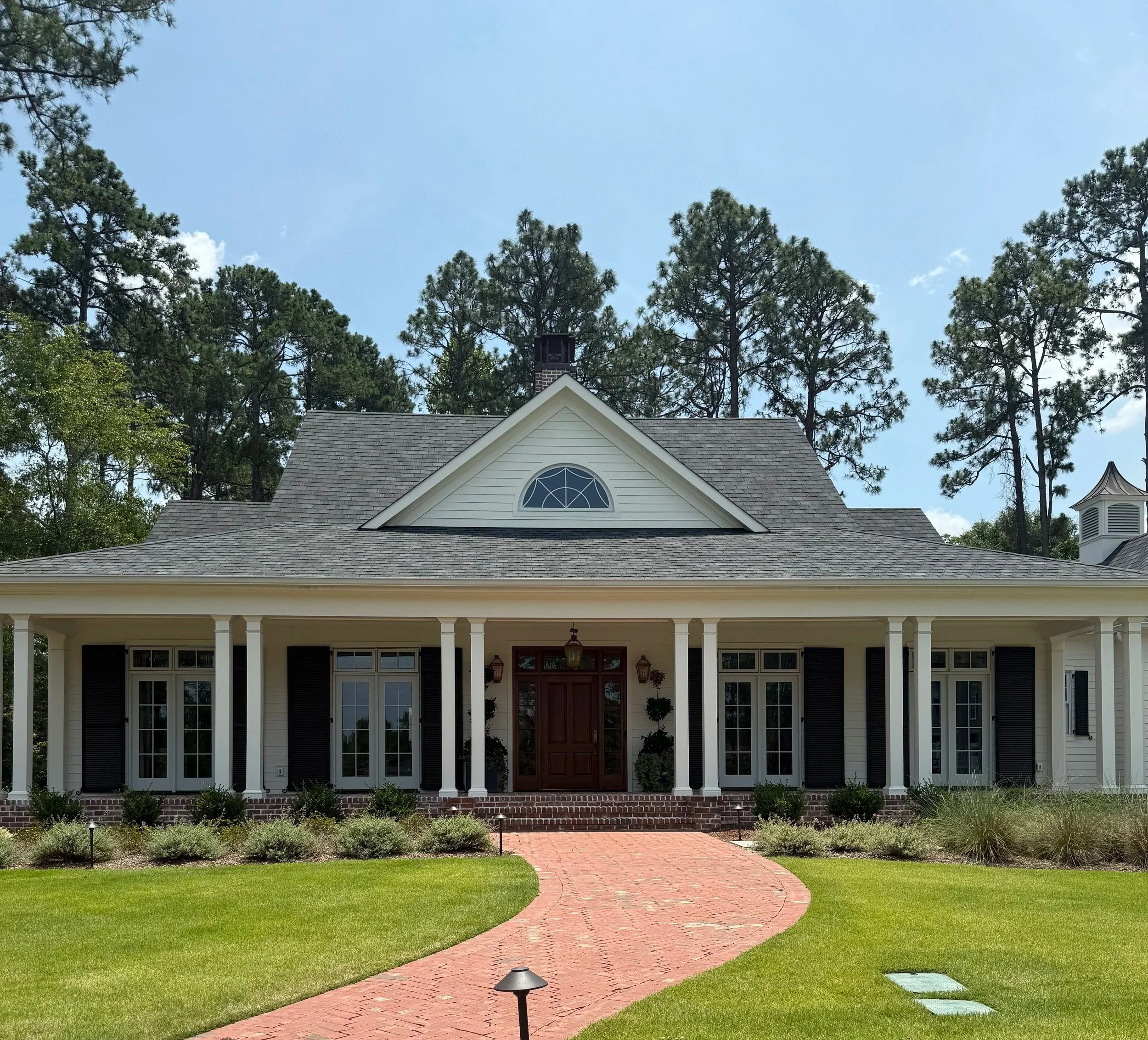Front view of a house with a gabled roof, porch with white columns, and a brick pathway leading to the front door, surrounded by green grass and trees.