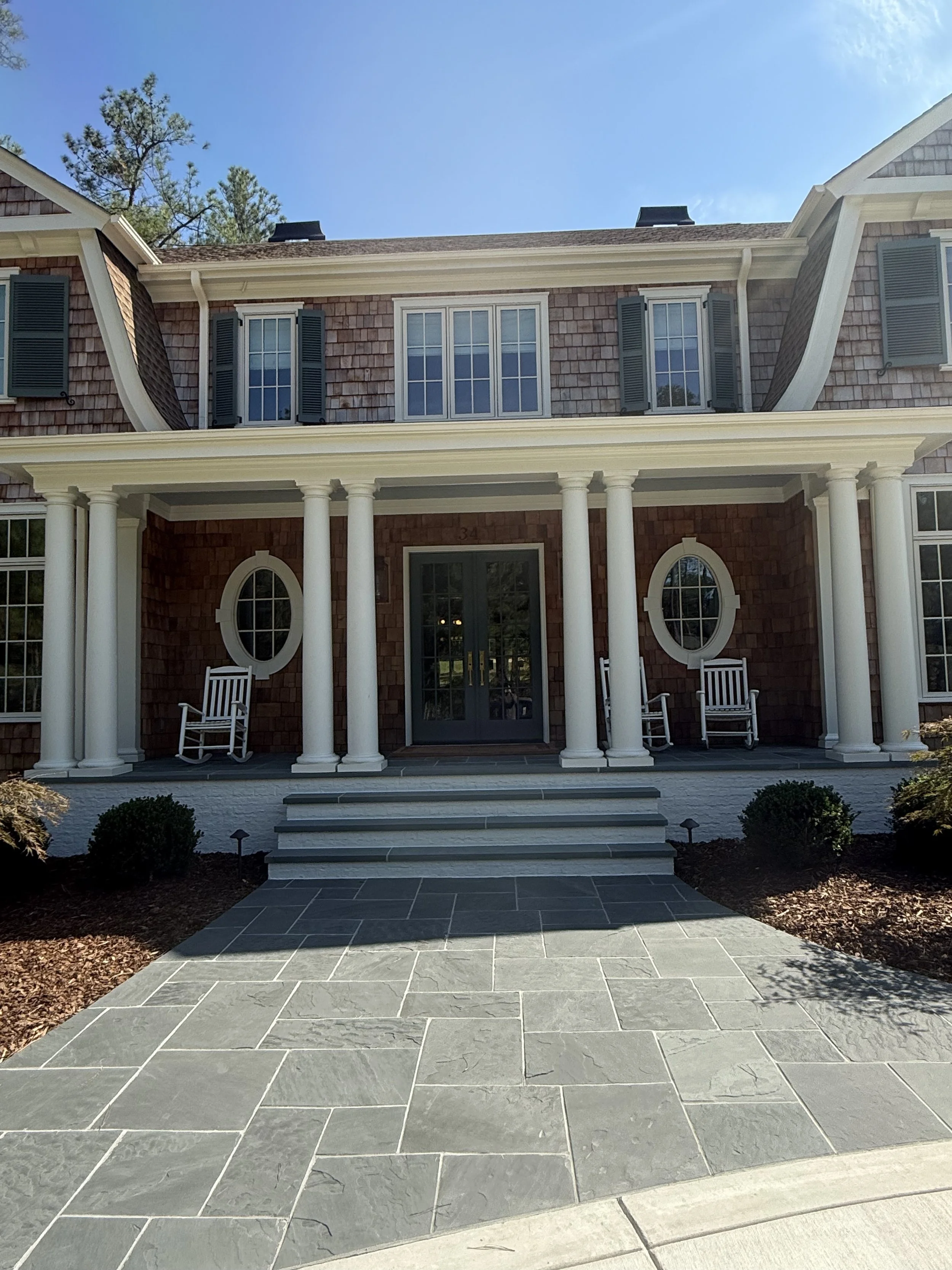 Front view of a house with brick exterior, white columns, steps leading to a door, two rocking chairs, and surrounding bushes, under a blue sky.