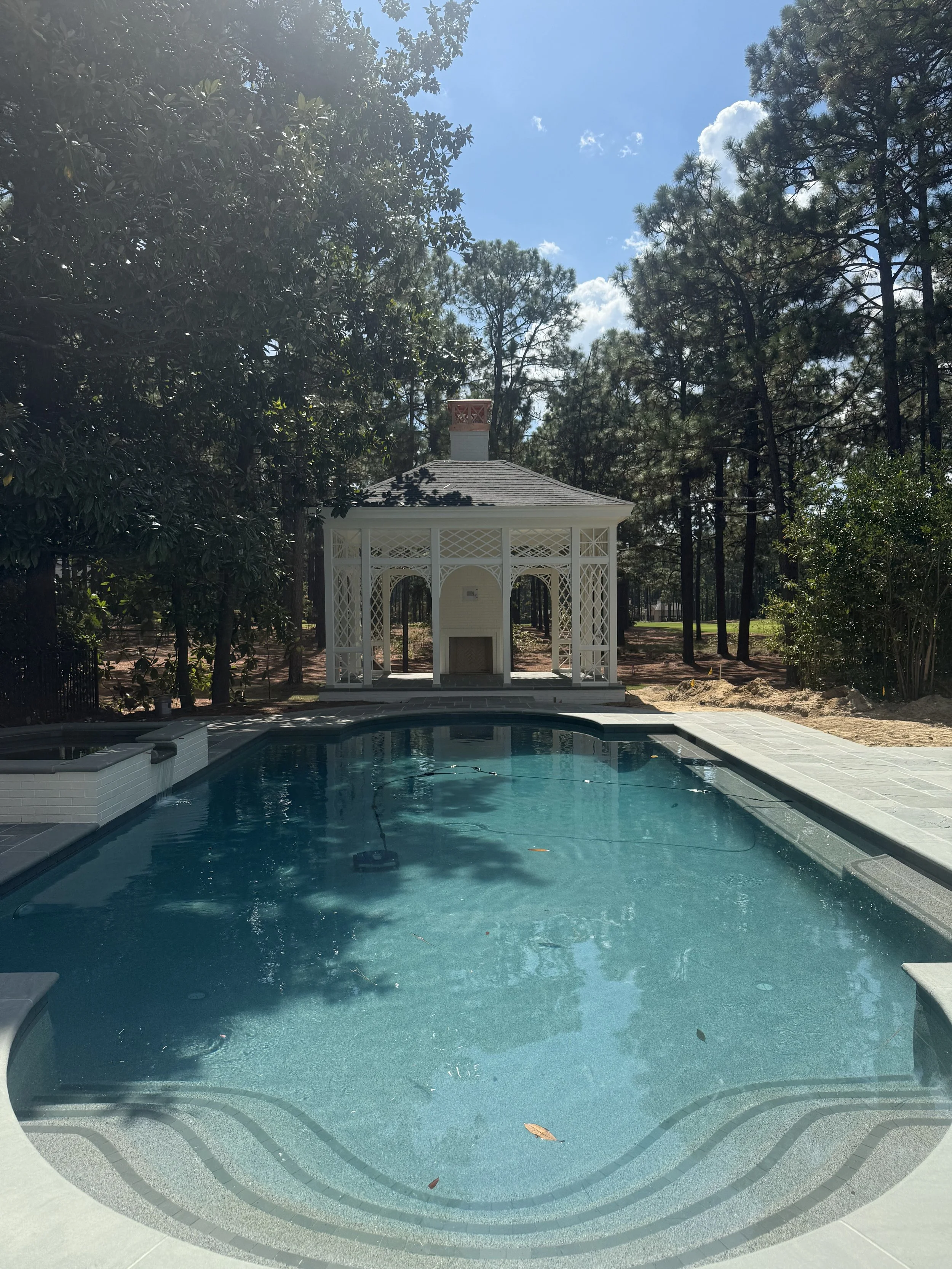 Swiming pool surrounded by a concrete deck with a small white gazebo at the background and tall trees on a sunny day.