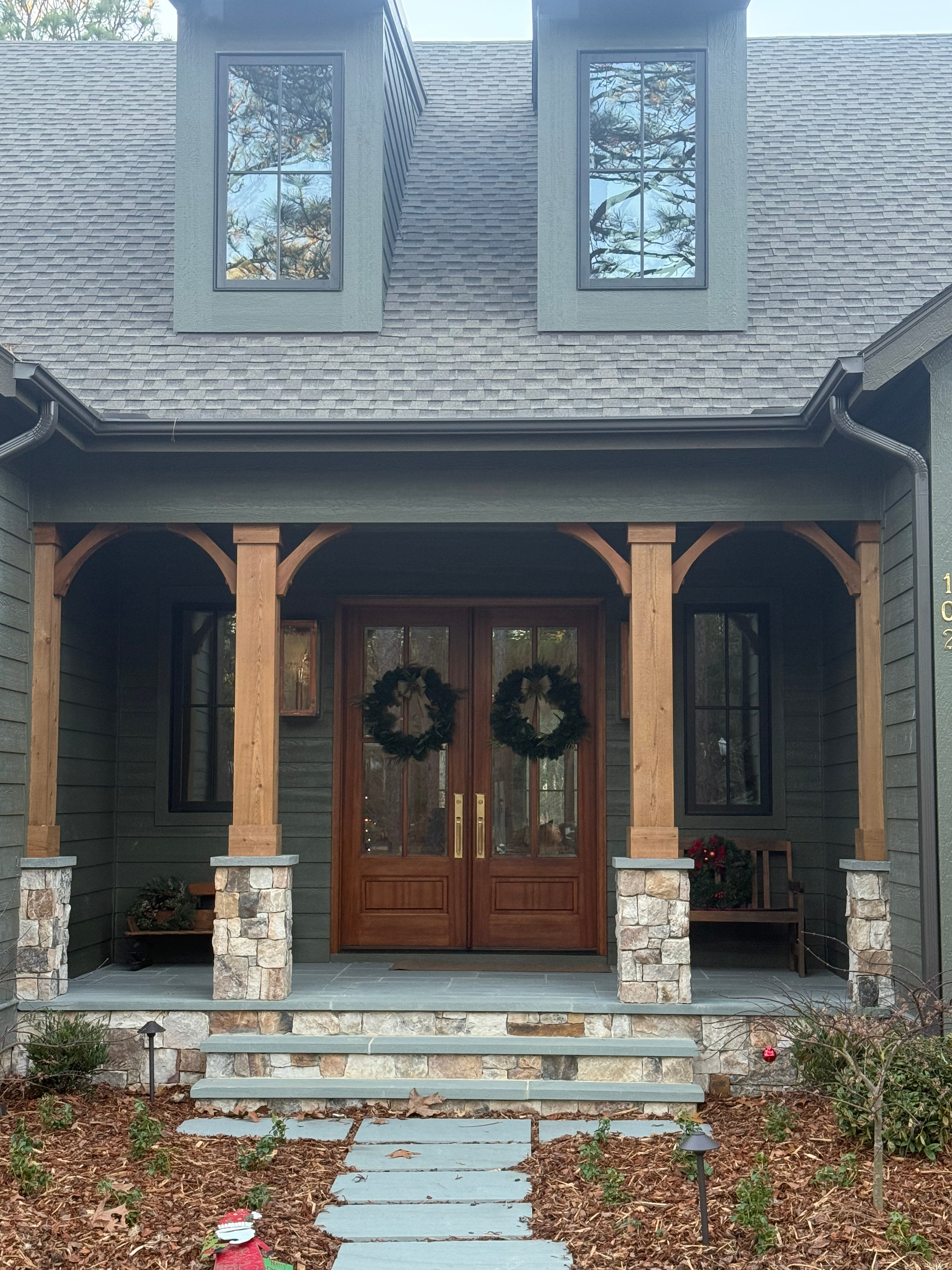 Front view of a house with a stone porch, wooden front door, and two gable windows reflecting trees. There are steps leading up to the porch, outdoor lights, and some small plants and decorations in the yard.