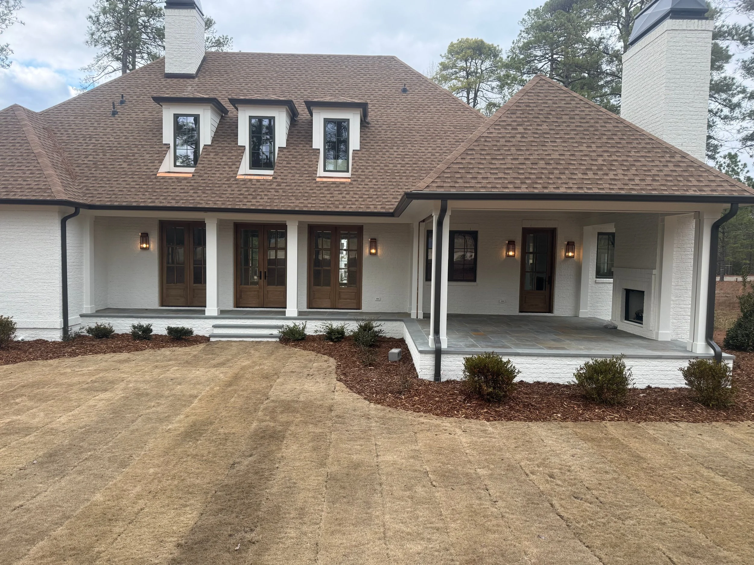 A modern house with a brown shingled roof, white brick walls, and wooden double doors at the entrance. There are three dormer windows on the roof and large chimney structures on both sides. The house has a covered porch area with outdoor lighting. Th