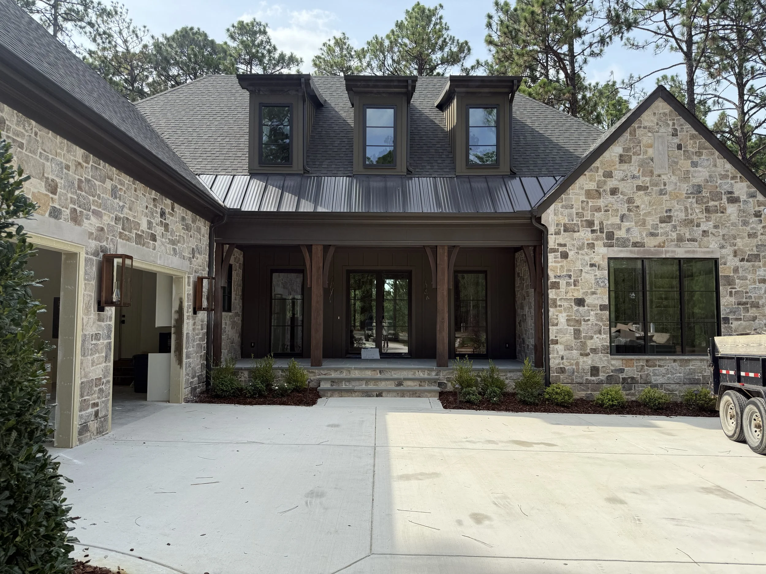 View of a house's backyard with stone walls, large windows, a sliding glass door, a covered porch with wooden beams, a paved concrete patio, and trees in the background.