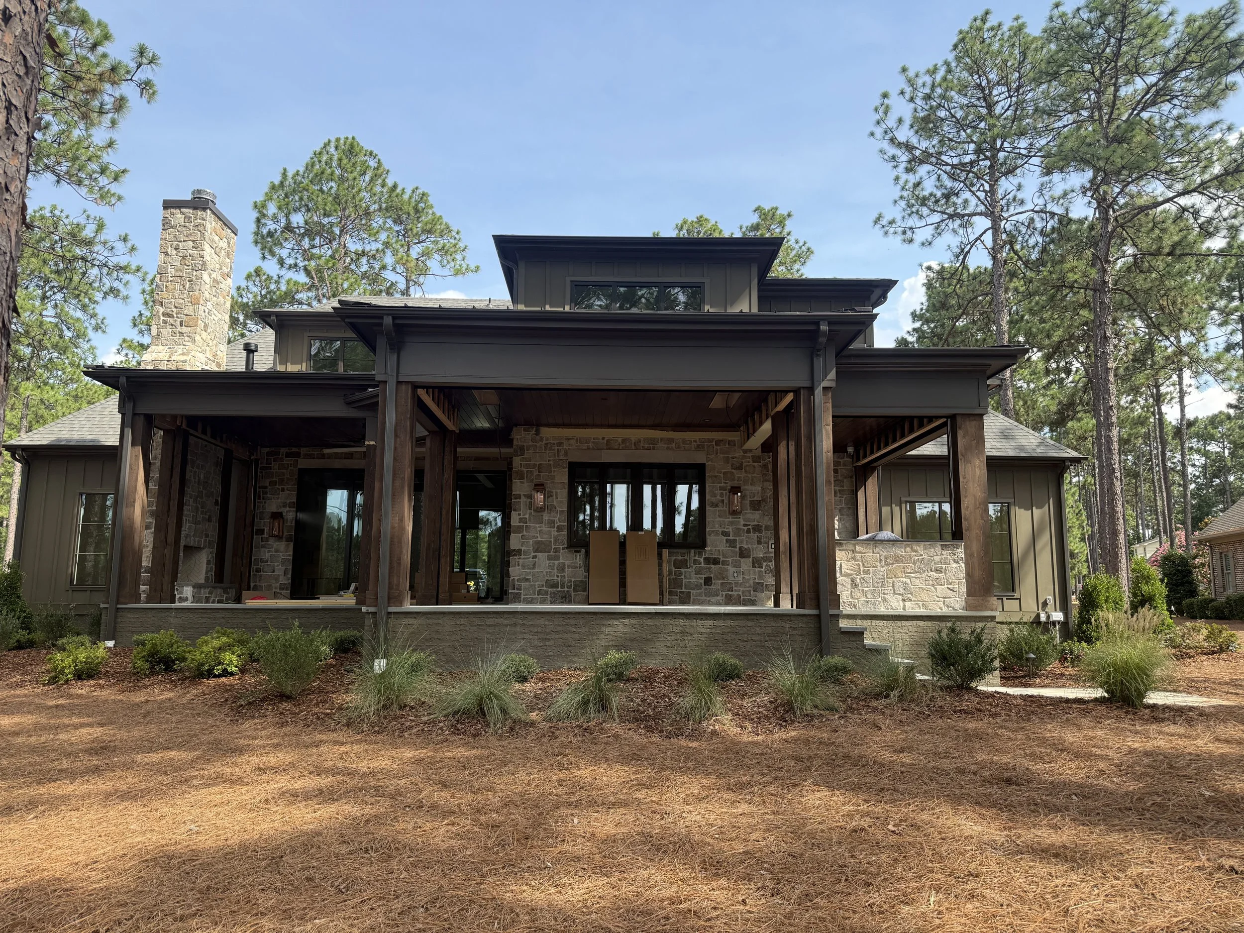 The back of a house with a wooden deck, stone and siding exterior, large windows, a chimney, surrounded by pine trees, and a landscaped yard with grass and bushes.