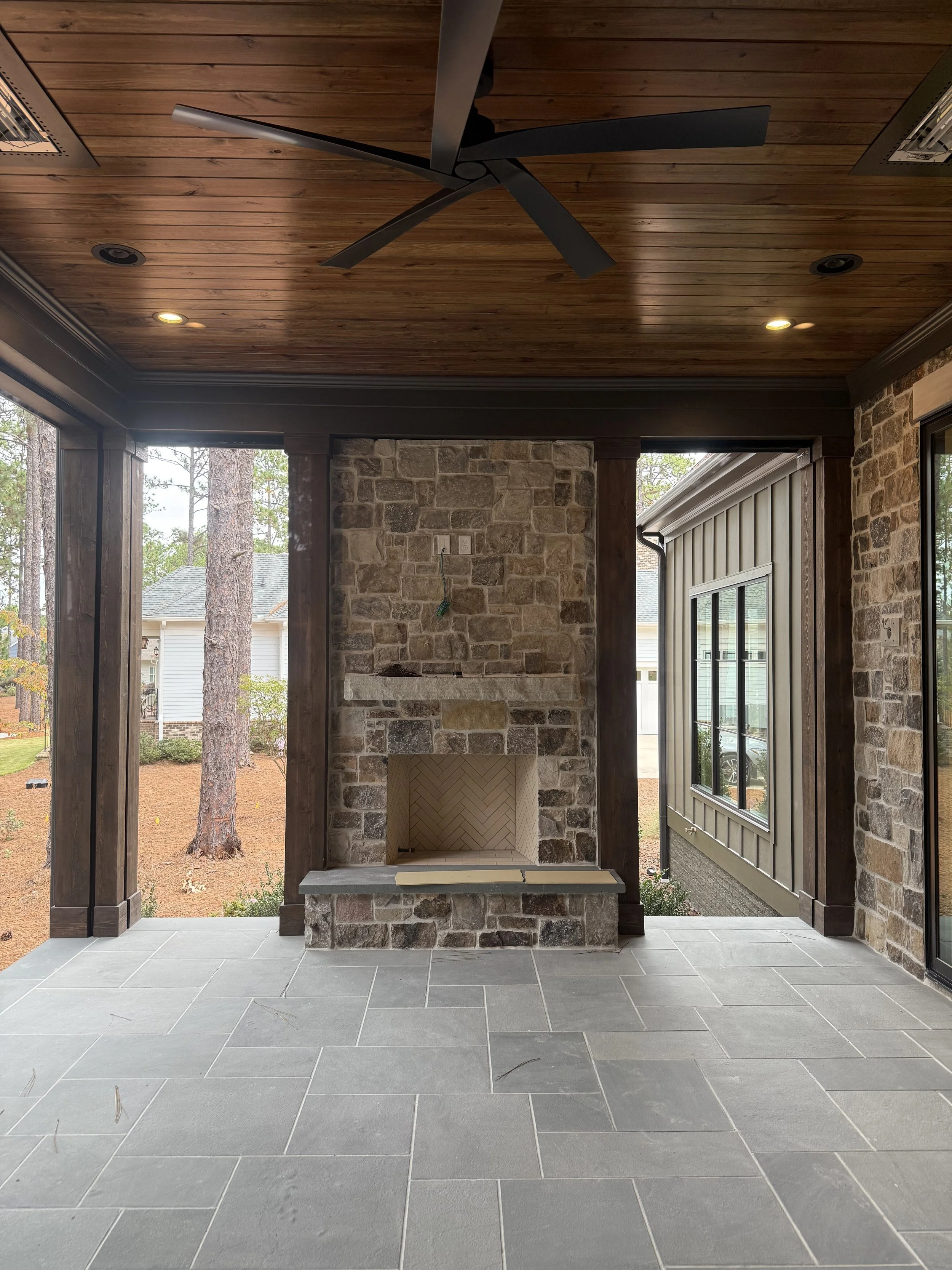 Interior view of a porch with a stone fireplace, wooden ceiling with recessed lighting, large sliding glass door, and tiled flooring, overlooking a yard with trees and neighboring houses.