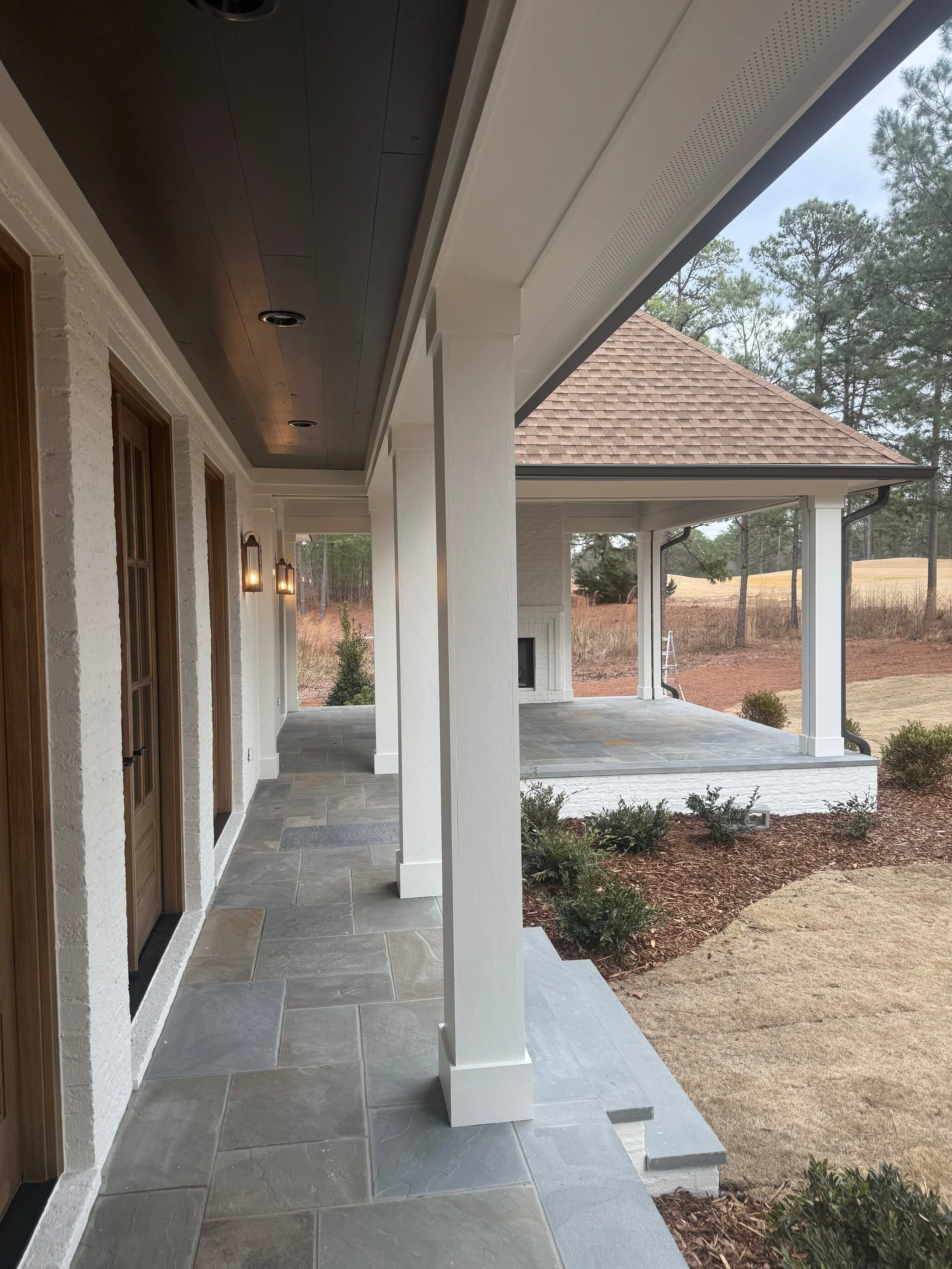Covered porch with stone-tile flooring, white columns, dark ceiling, outdoor wall-mounted lights, and a view of trees and a sloped yard.