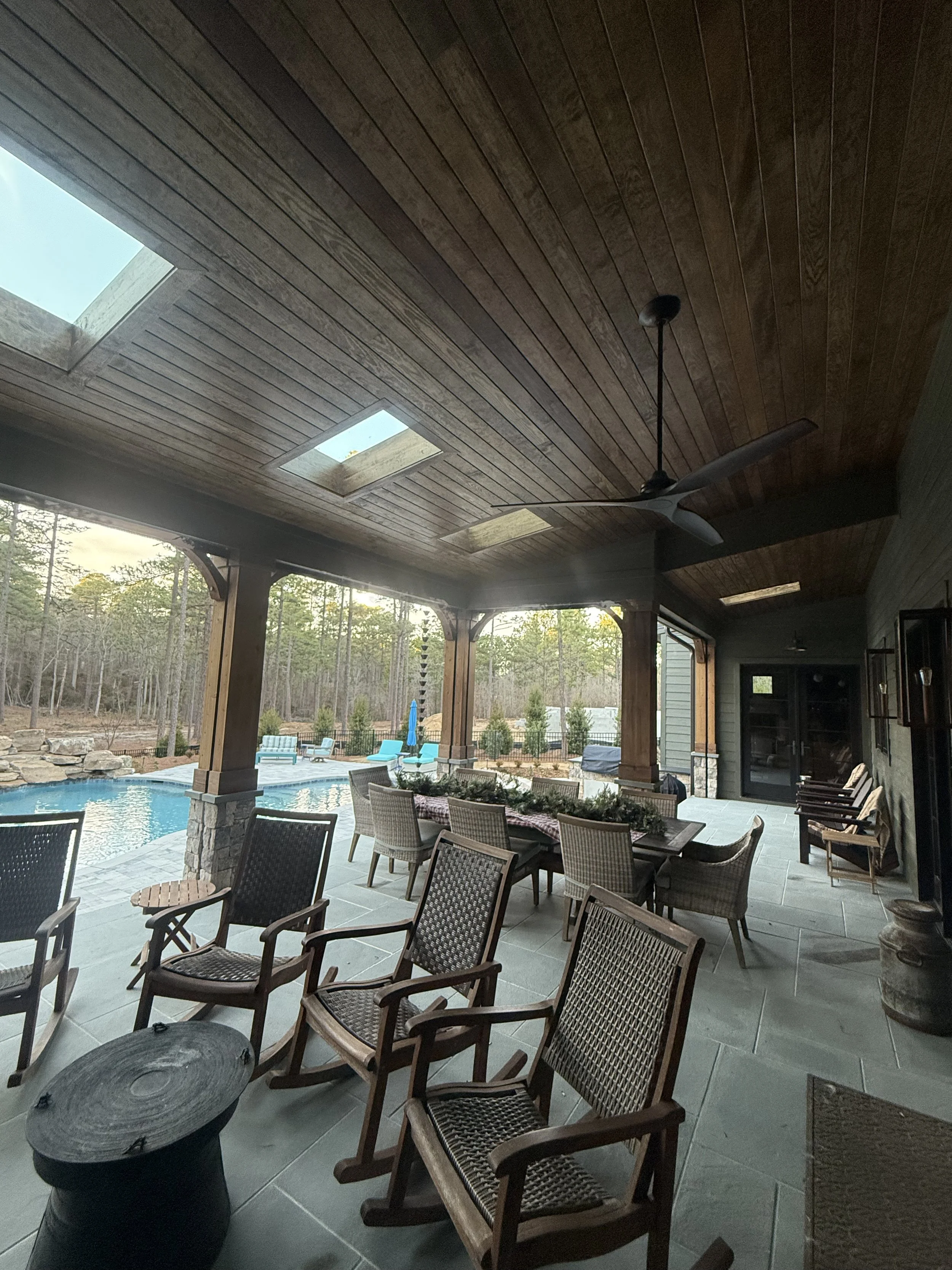 Covered patio area with outdoor dining table, multiple chairs, ceiling fan, and views of a backyard with swimming pool, outdoor furniture, and trees.
