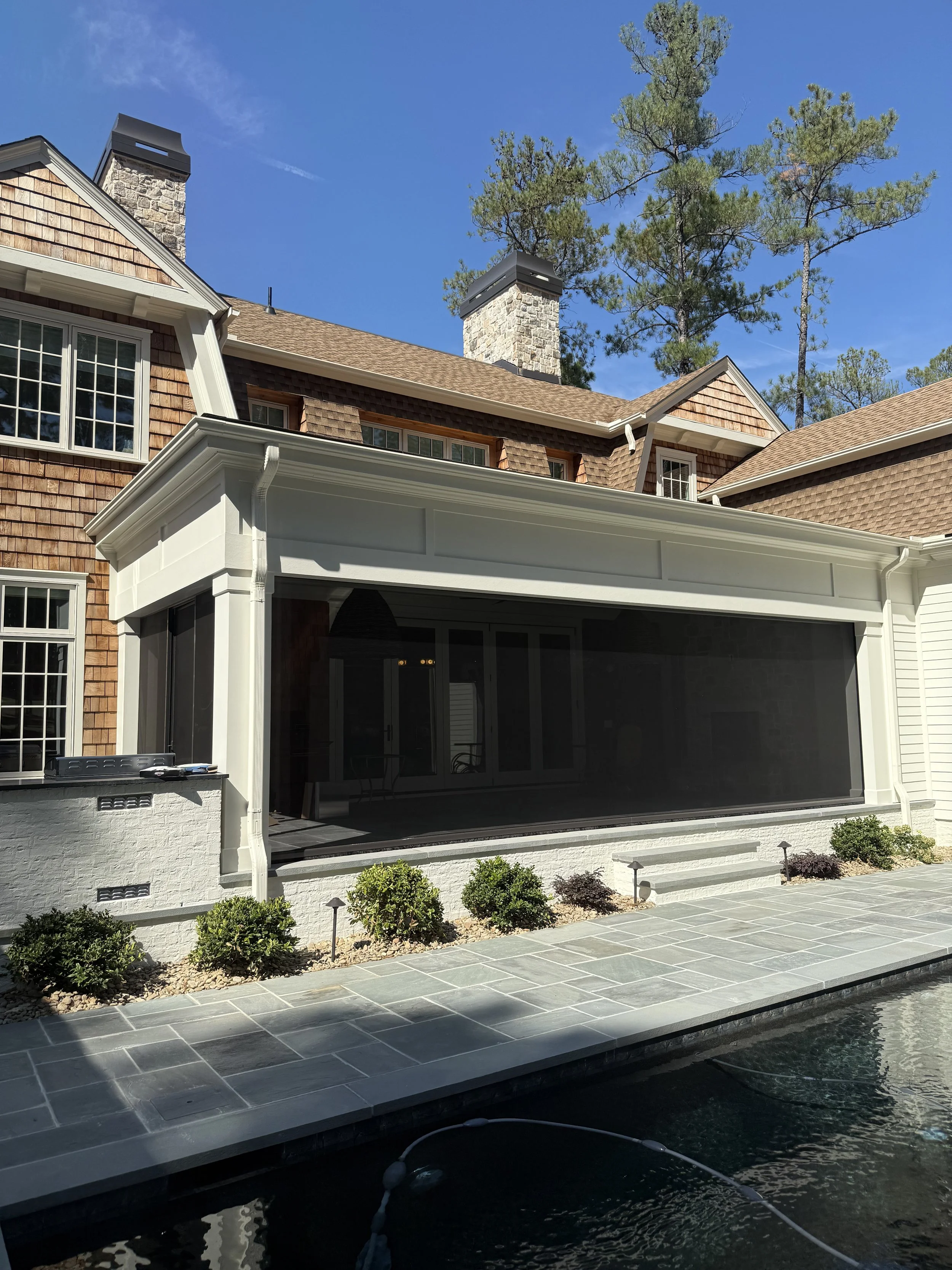 View of a house with a screened porch, stone chimney, and shingles on the roof. There are trees in the background and a pool with a tiled edge in the foreground.