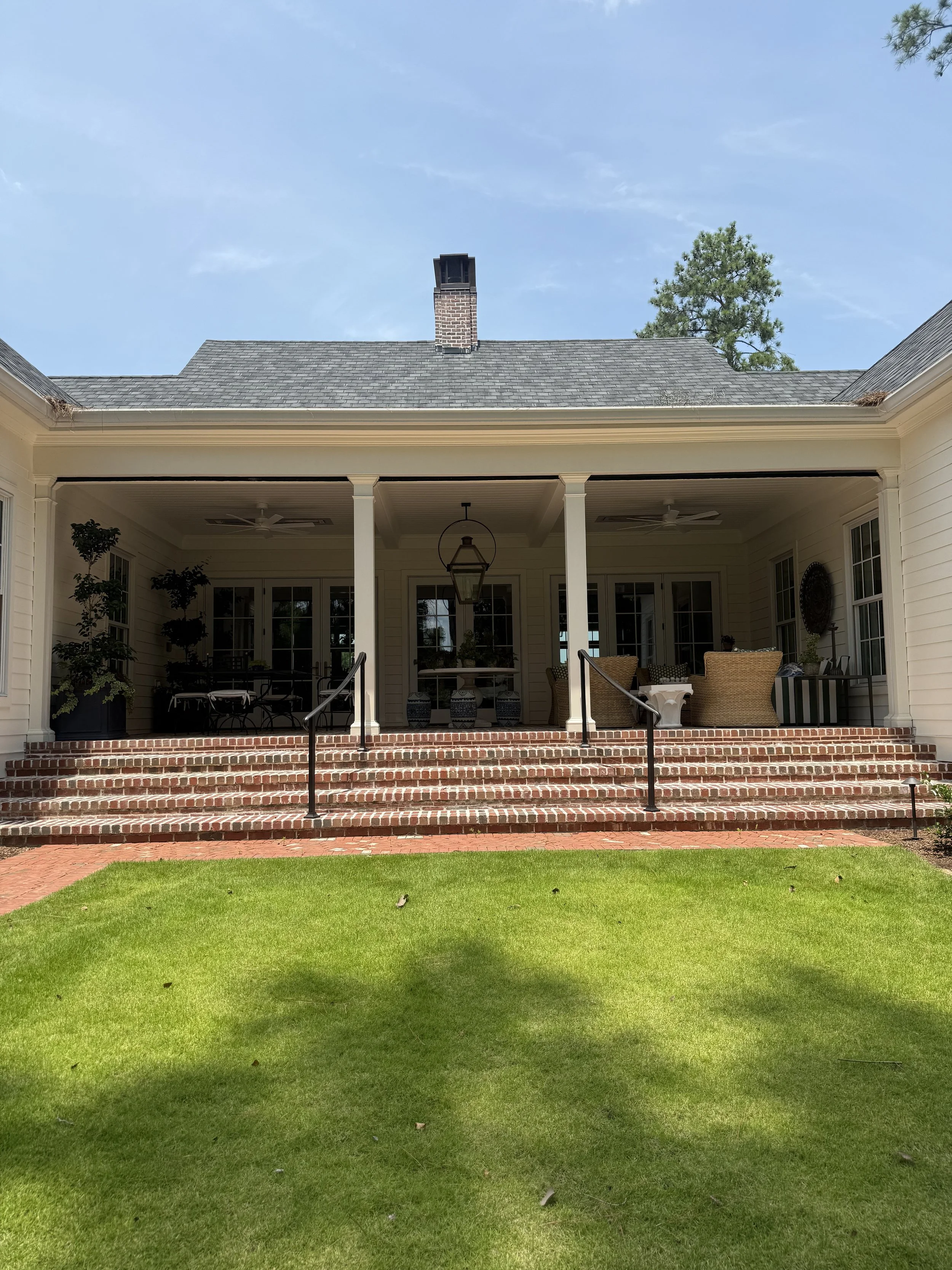 Back view of a house with a covered porch, brick steps, and outdoor furniture, overlooking a green lawn under a blue sky with a few clouds.