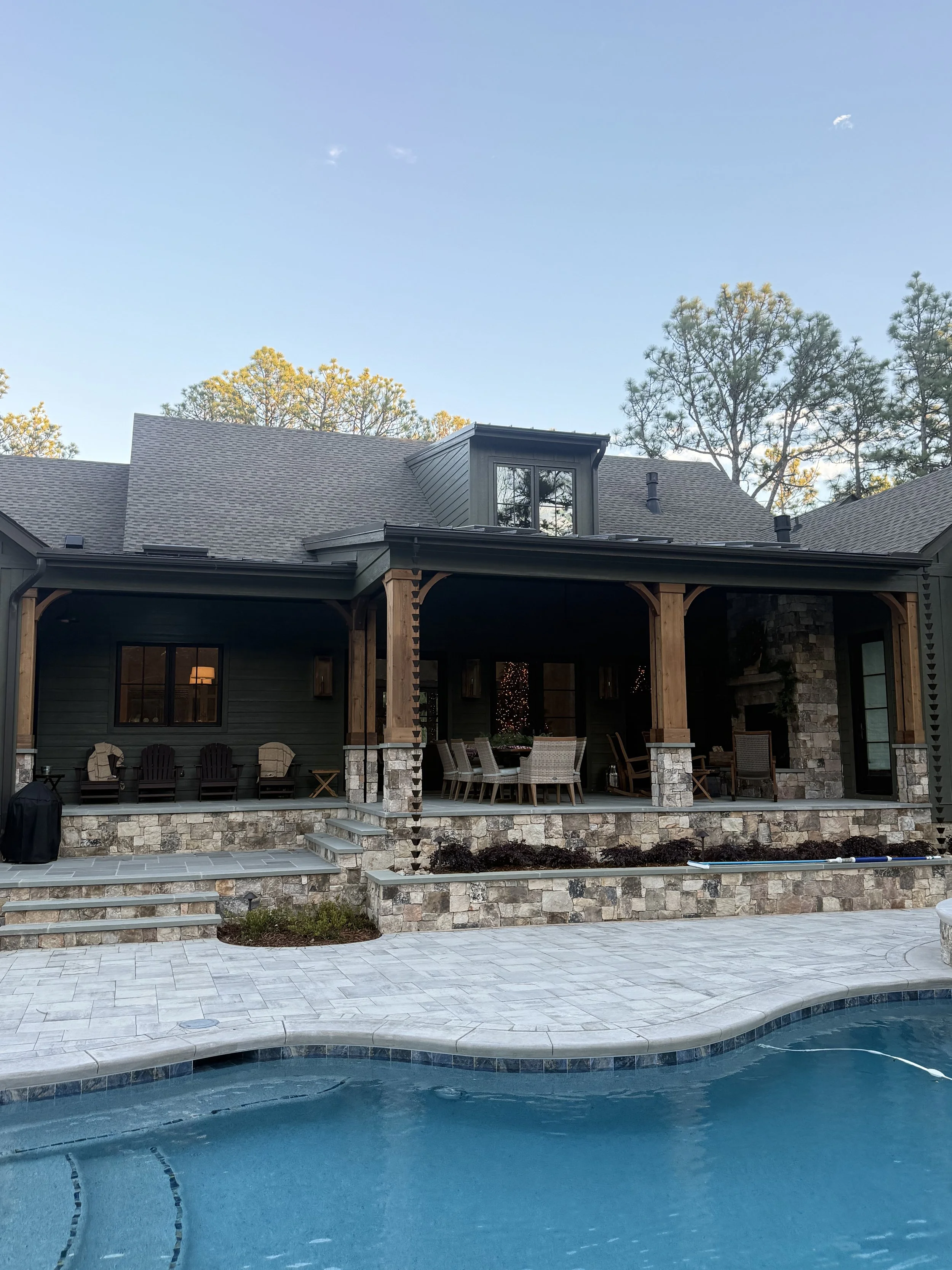Backyard view with a swimming pool, patio area with chairs and table, house with stone and dark siding exterior, and trees in the background under a clear blue sky.