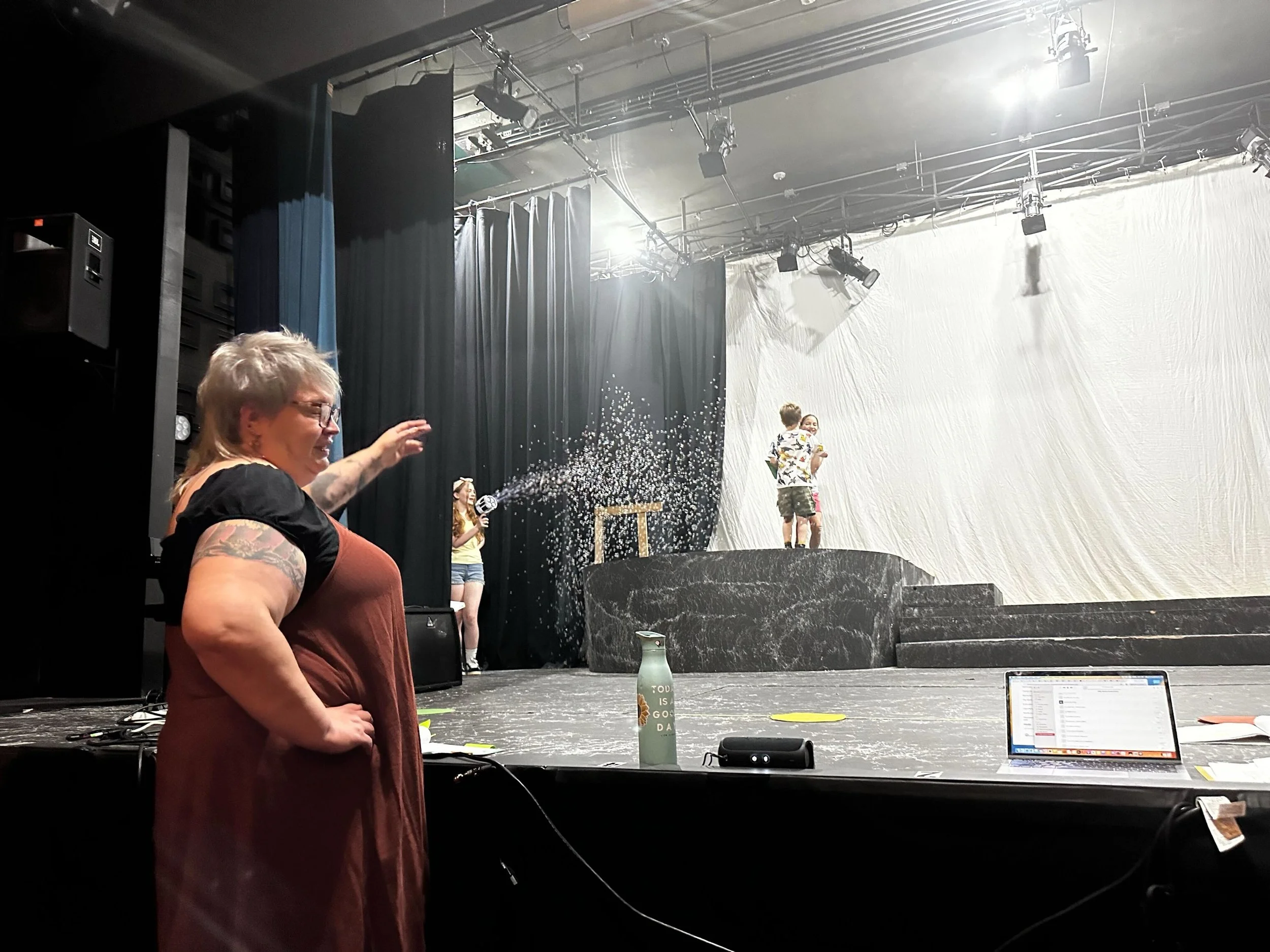 a woamn in the foreground wearing an orange jumpsuit is pointing to kids onstage. One is holding a bubble machine and bubbles are blowing on stage.