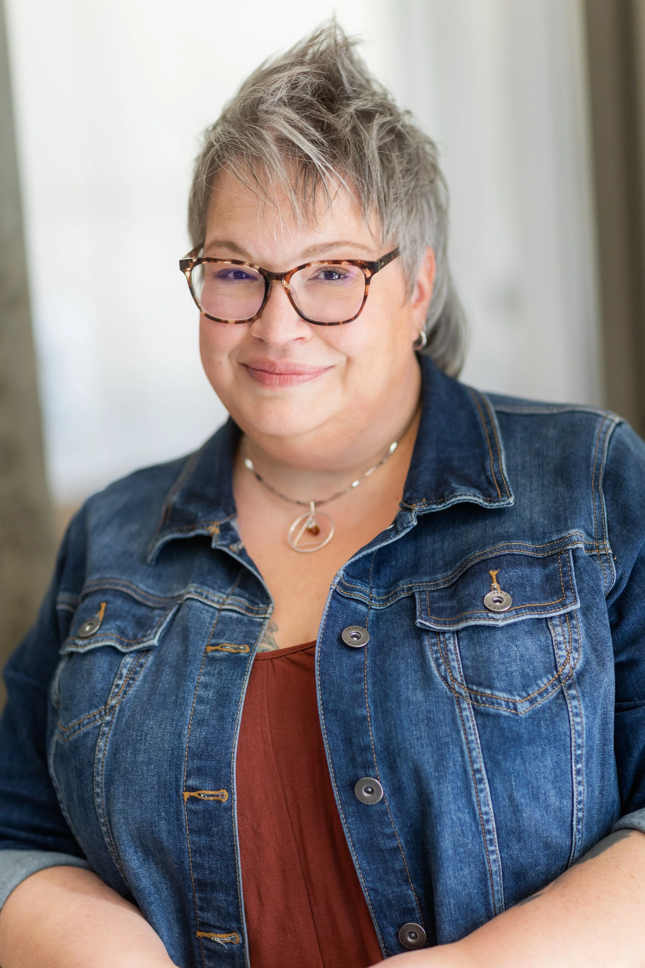 A woman with short spiky blonde hair smiles at the camera. She's wearing glasses, a red shirt, and a jean jacket.