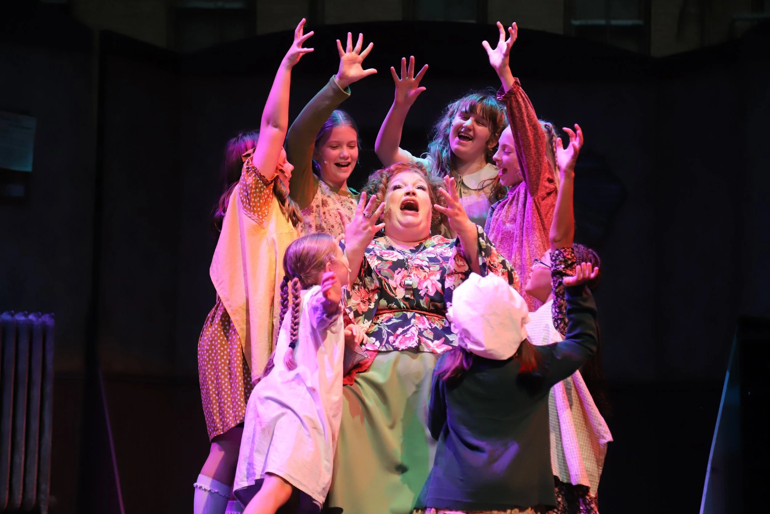 a group of young girls with their arms up surround an angry woman who is sitting down