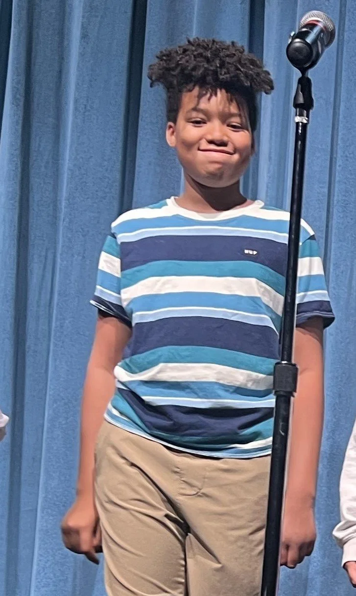 A young child with a blue and white striped shirt stands in front of a blue curtain near a microphone.