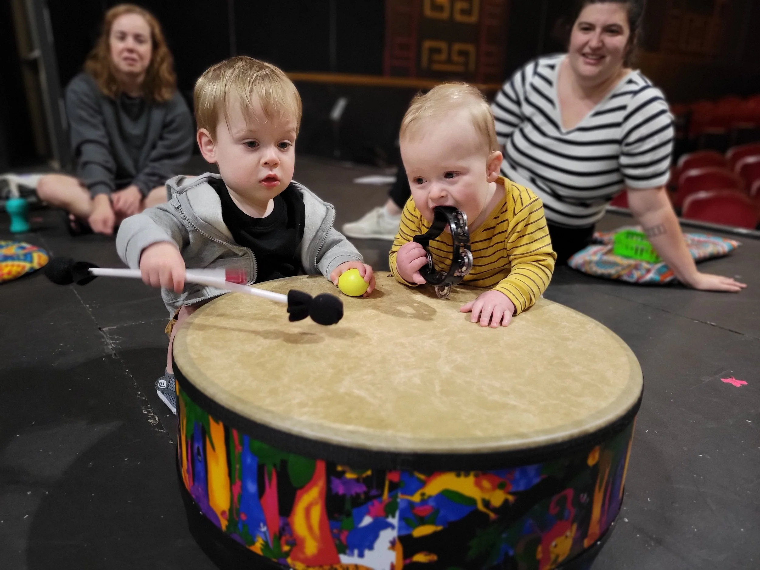 Two young boys playing on a colorful drum, one holding a yellow ball and the other biting on a tambourine, with two women sitting in the background observing them.