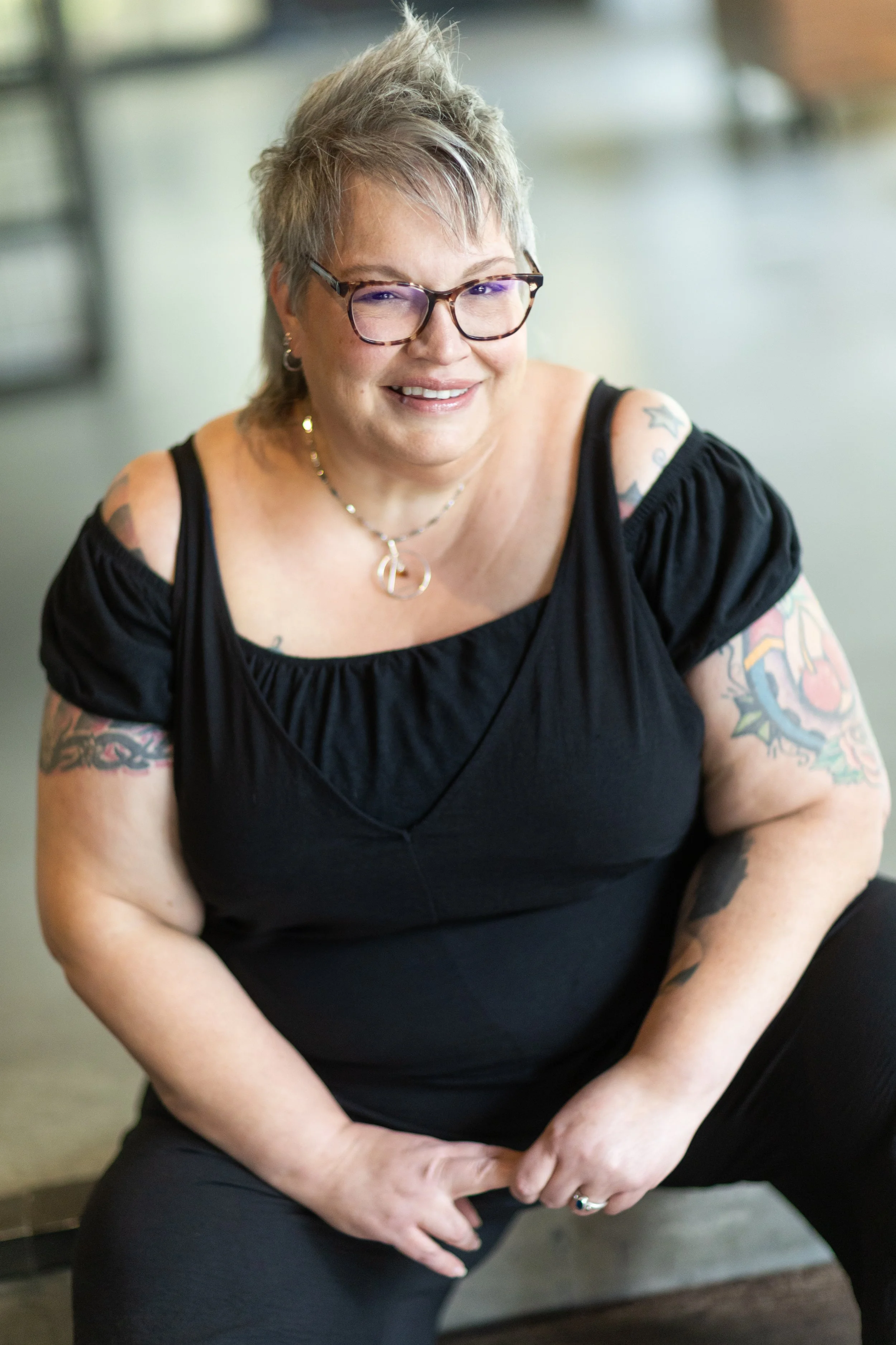 A person with short blonde hair and purple glasses smiles at the camera. She's wearing a black shirt and has tattoos on her arms.