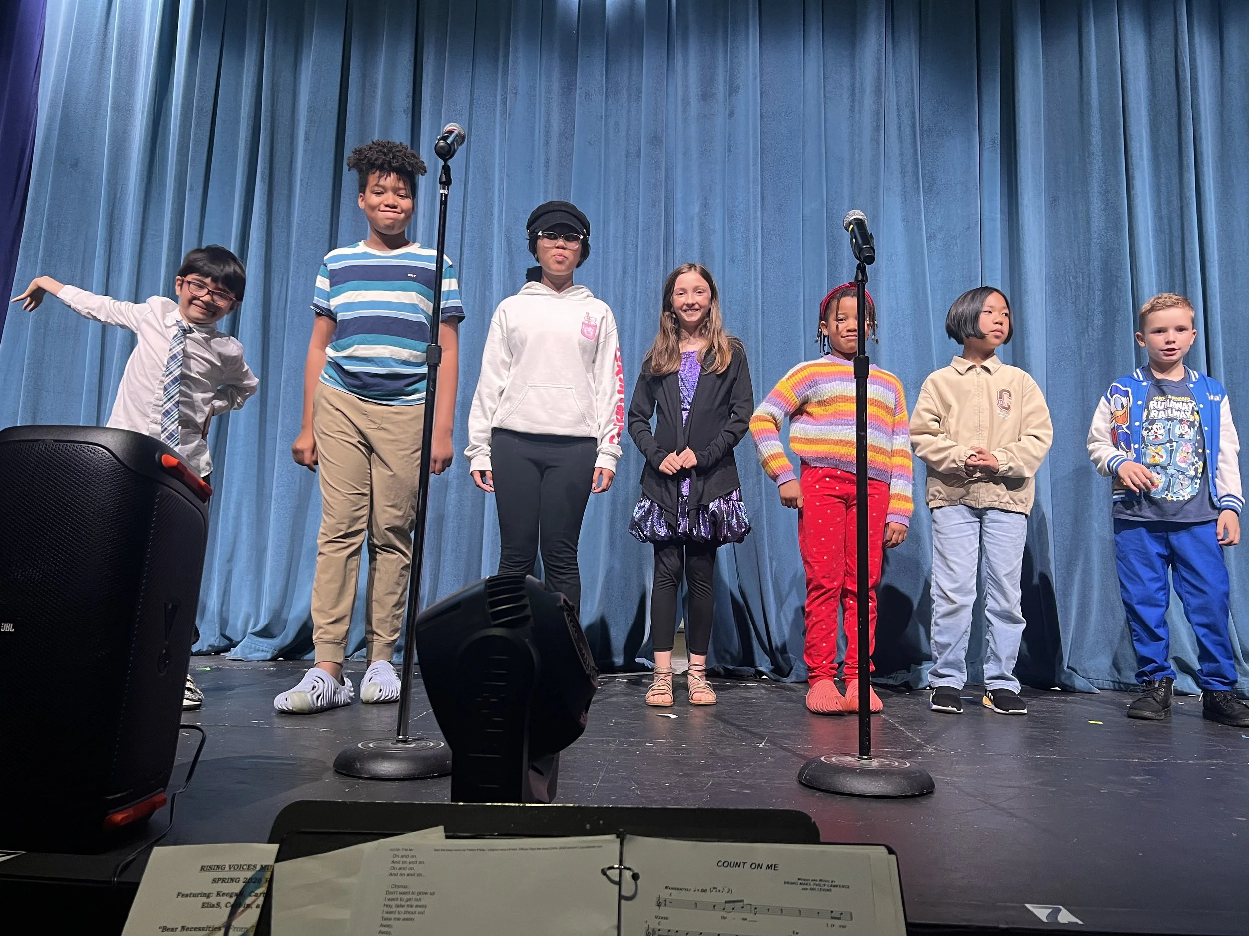 A group of young kids stands onstage in front of a blue curtain with microphones in the foreground.