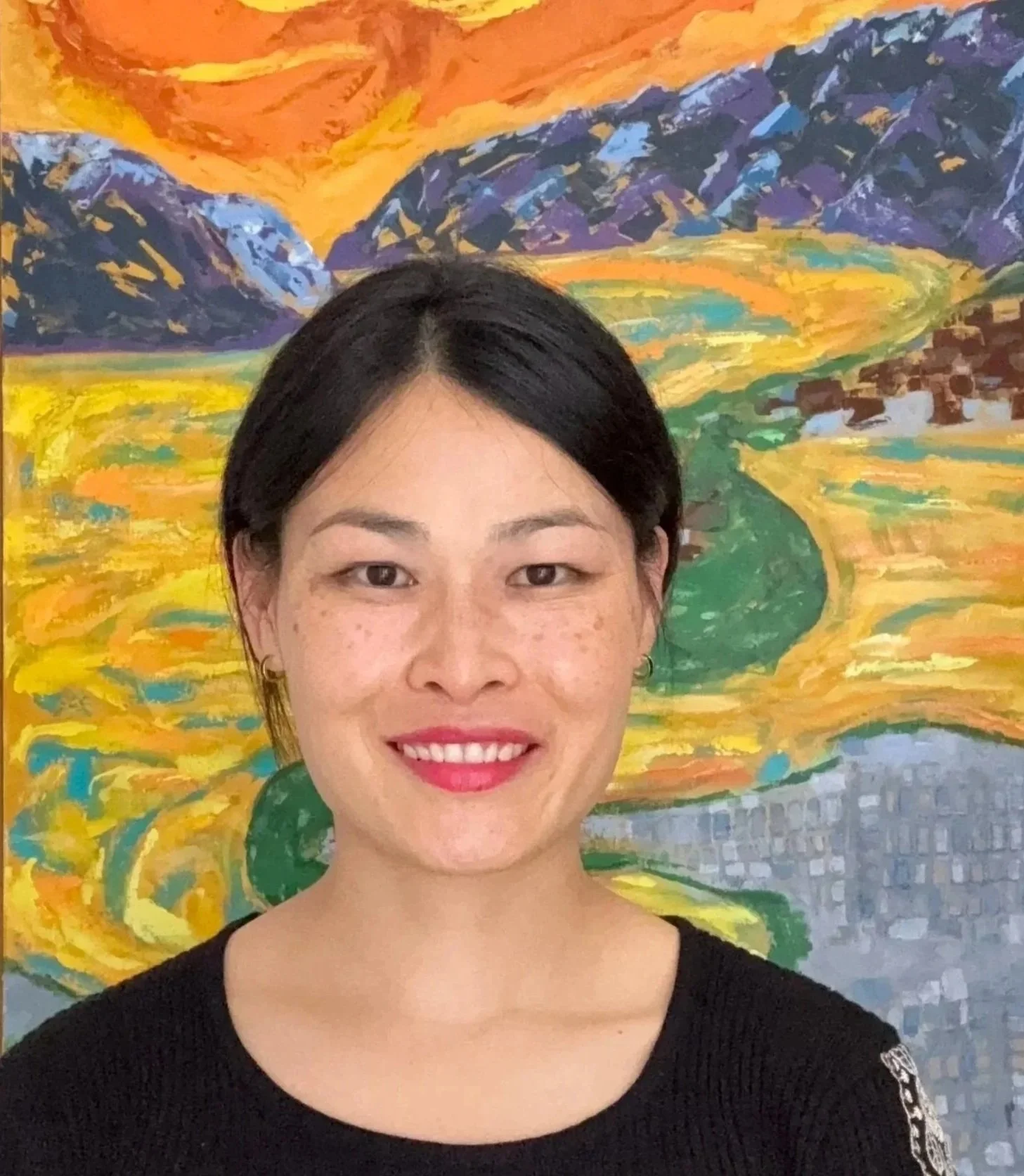 Cher Hou standing in front of her colourful Queenstown-inspired landscape painting featuring mountains, water and a vibrant sky.