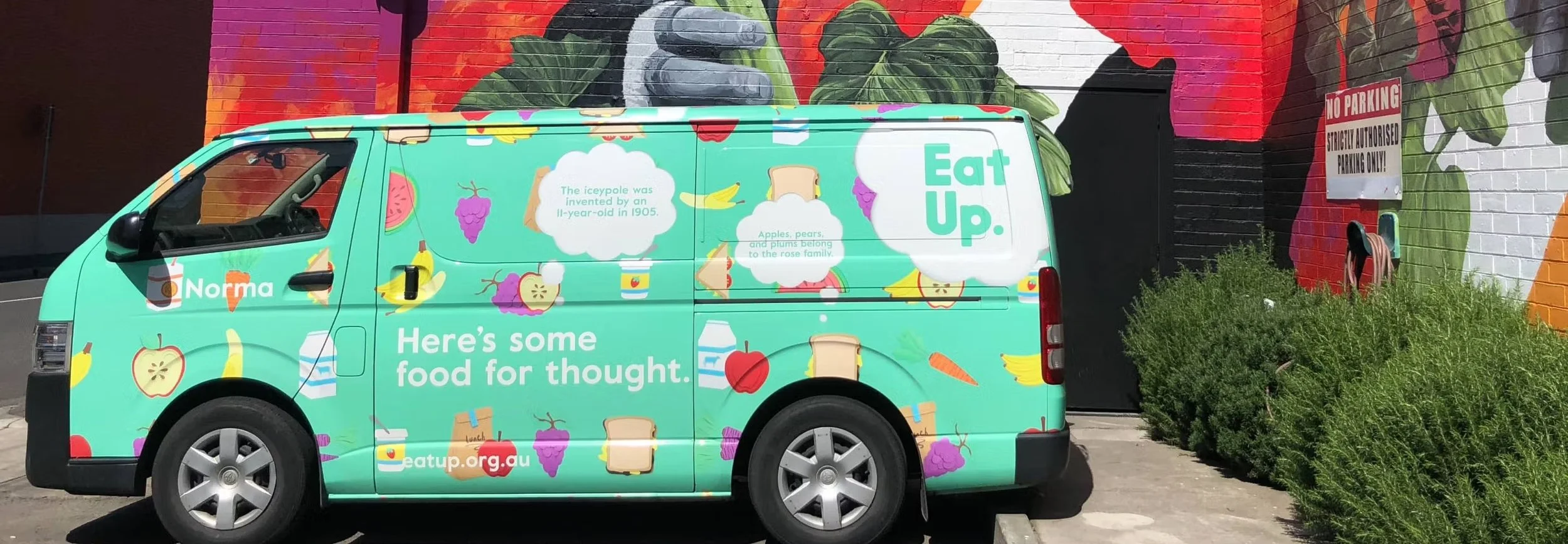 A teal food truck with colorful fruit and bread illustrations, parked in front of a mural with large leaves and a No Parking sign.