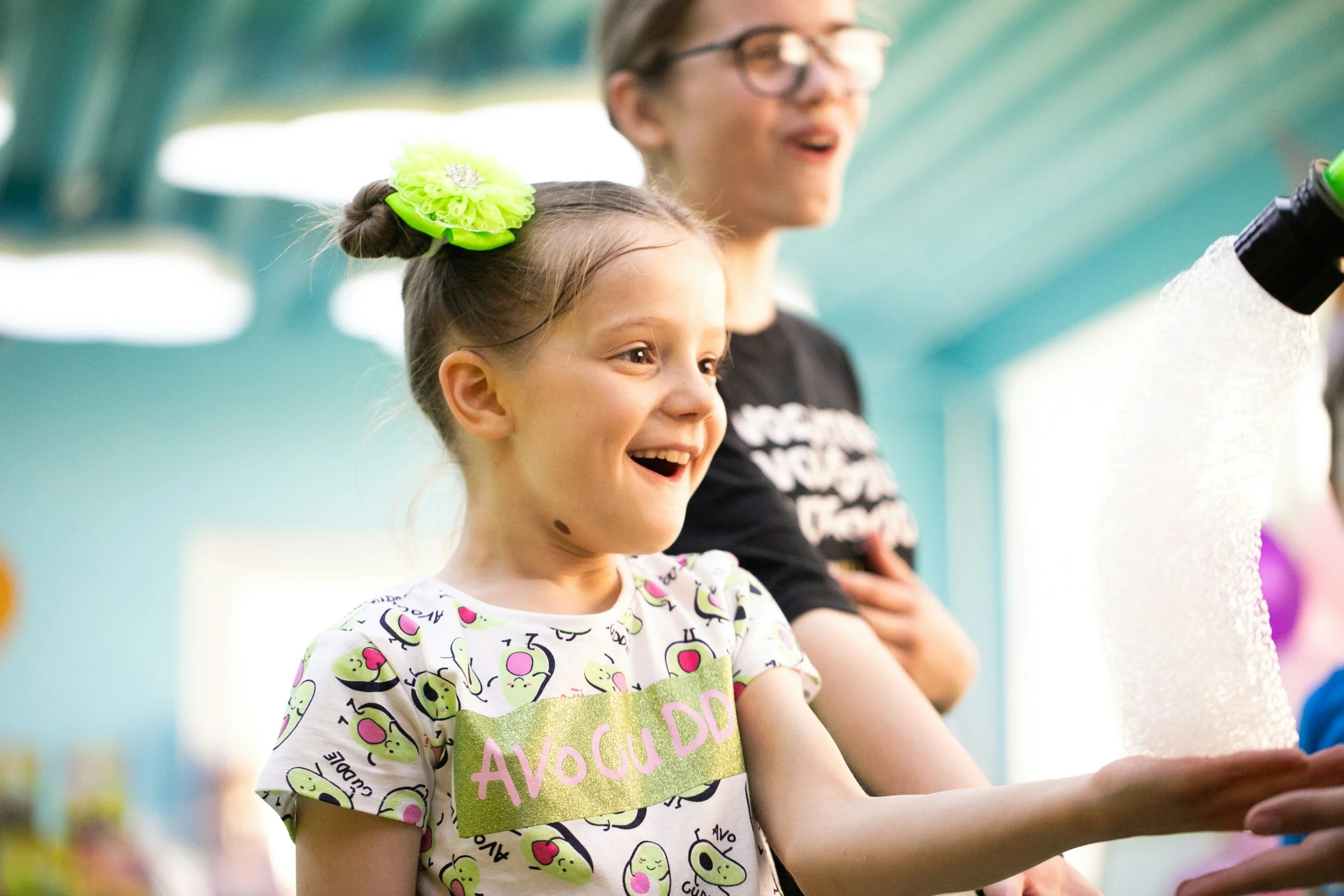 A young girl with a big green hair bow, smiling and wearing a avocado-themed shirt, standing with an older girl with glasses.