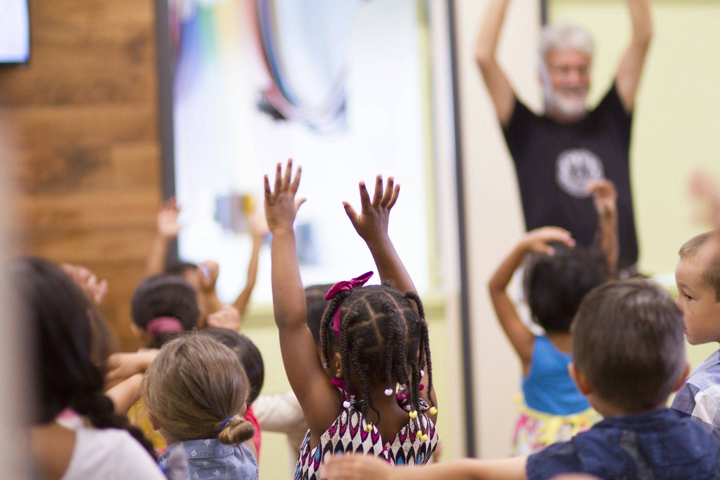 Children with hands raised during a classroom activity, with a teacher or adult in the background.