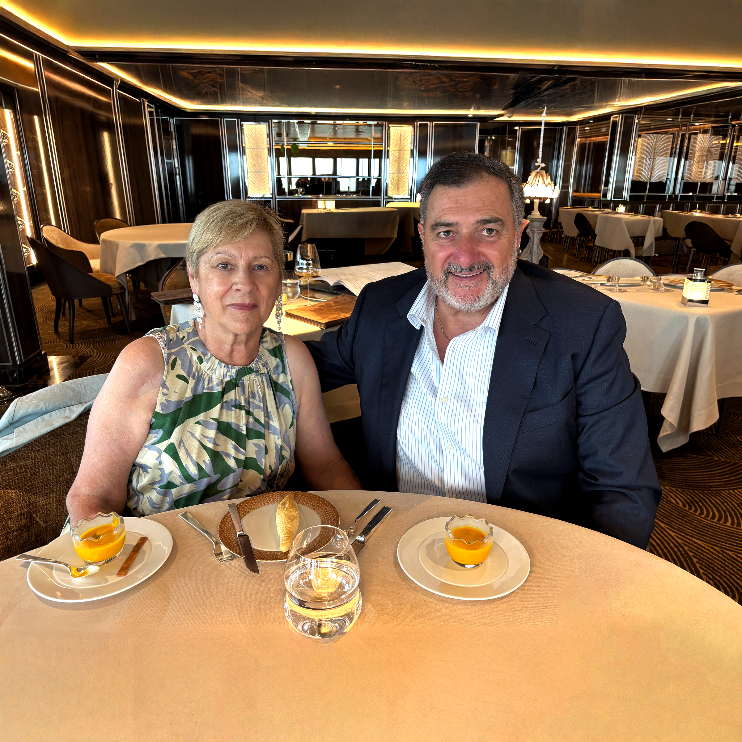 A woman and a man sitting at a restaurant table, smiling for a selfie. The table is set with menus, glasses, and small bowls of soup or sauce. The restaurant has a modern, luxurious decor with reflective surfaces and warm lighting.