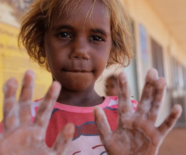 Close-up of a girl with messy blonde hair and brown skin showing her hands covered in mud or clay, with a blurred background of buildings and another child in the distance.