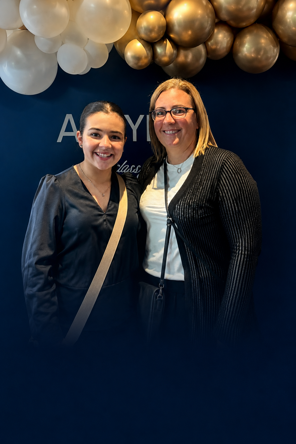 Two women standing together, smiling in front of a dark blue background with white and gold balloons overhead.