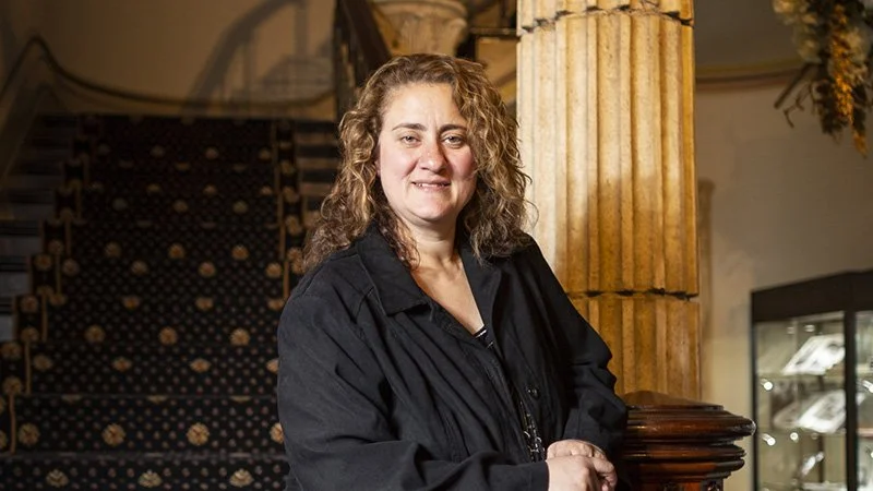 A woman with curly brown hair wearing a black jacket standing indoors near a decorative column and staircase.
