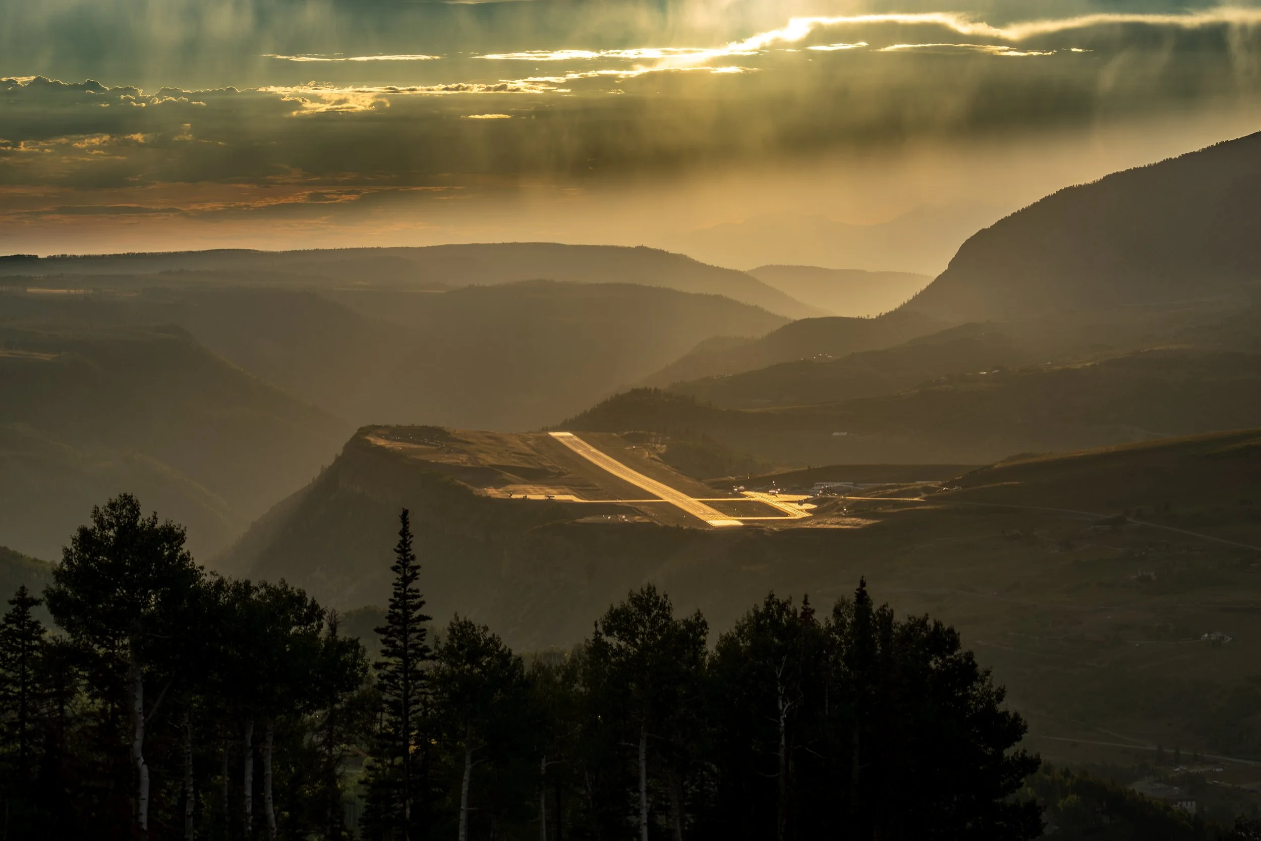 Hilly landscape with dense trees in foreground and winding road on hillside, mountains in background, with sunlight breaking through clouds.