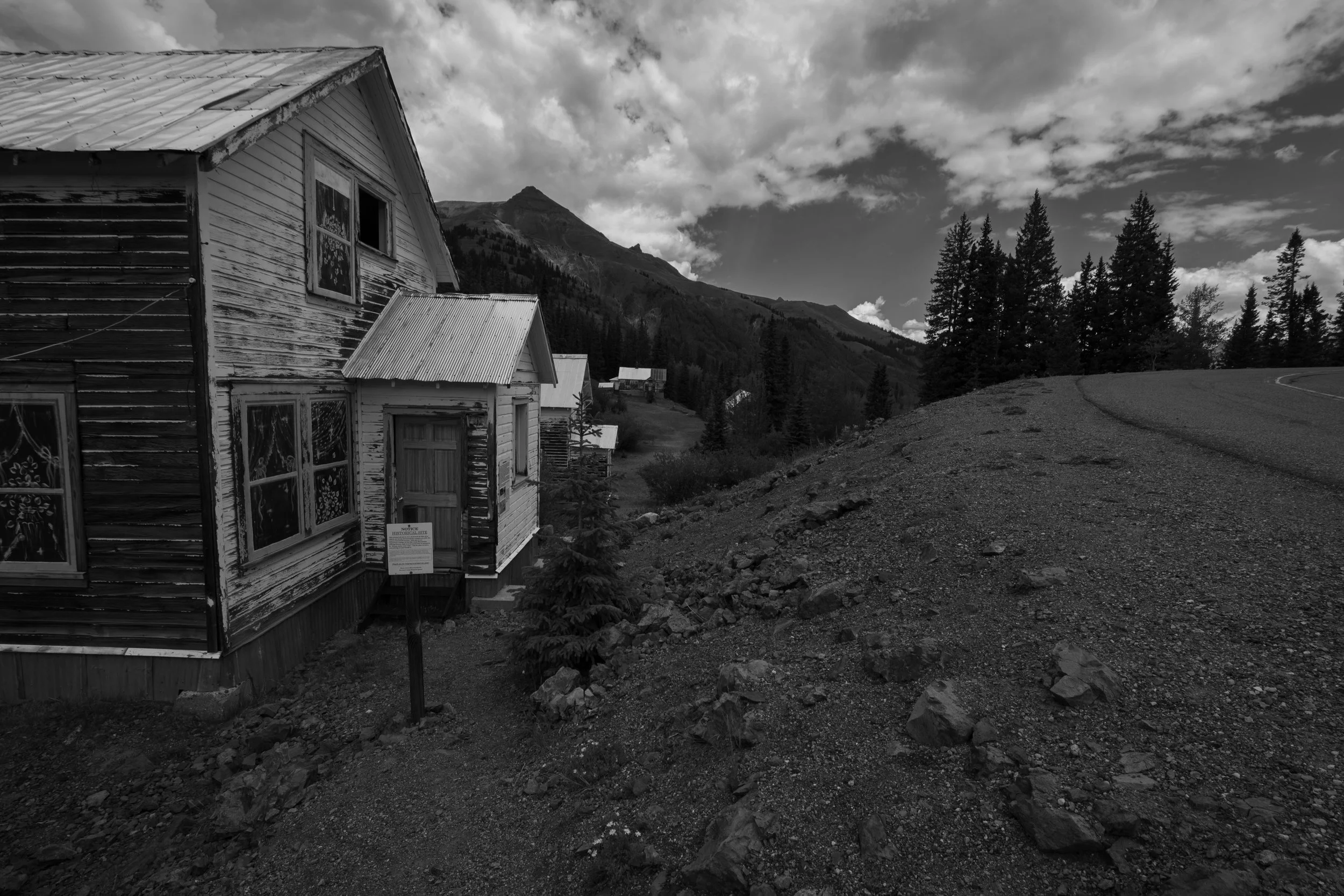 Black and white photo of old, weathered wooden houses with a mountain and pine trees in the background, under cloudy sky.