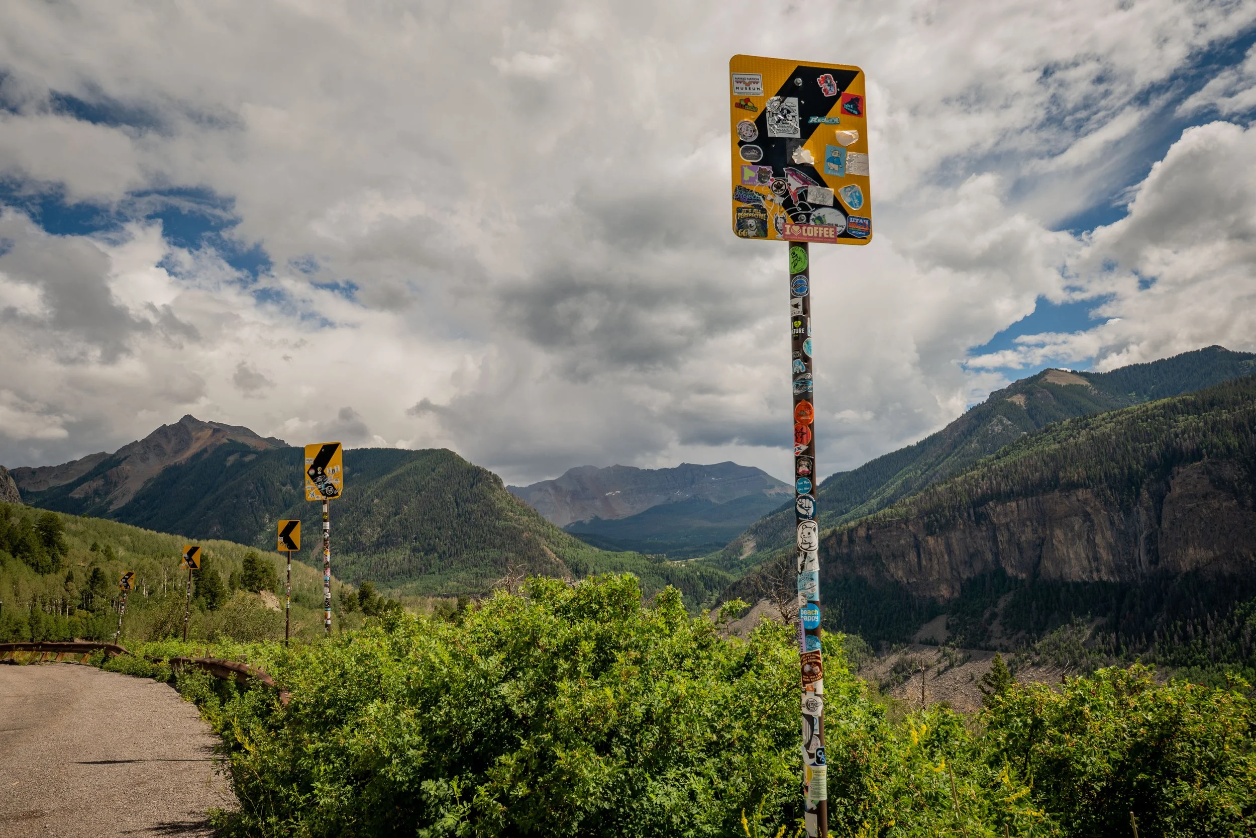 Tall pole covered with numerous stickers and patches, located alongside a mountain road with mountain peaks and cloudy sky in the background.