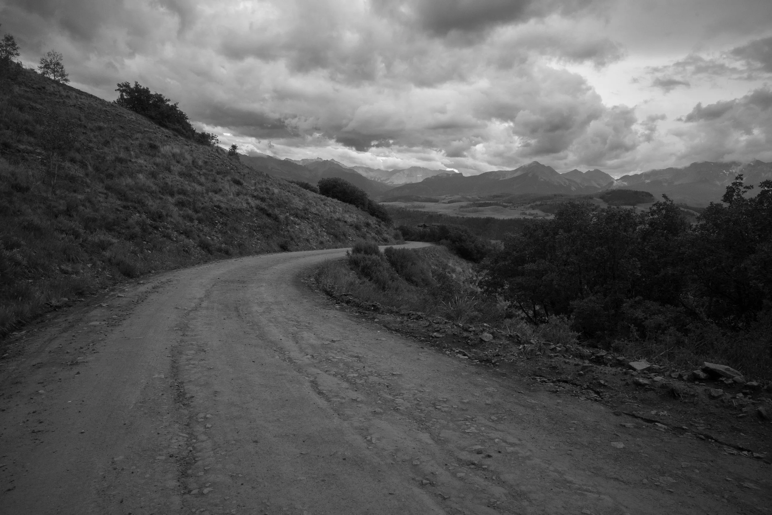 Black and white photo of a winding dirt road through mountainous landscape with cloudy sky.