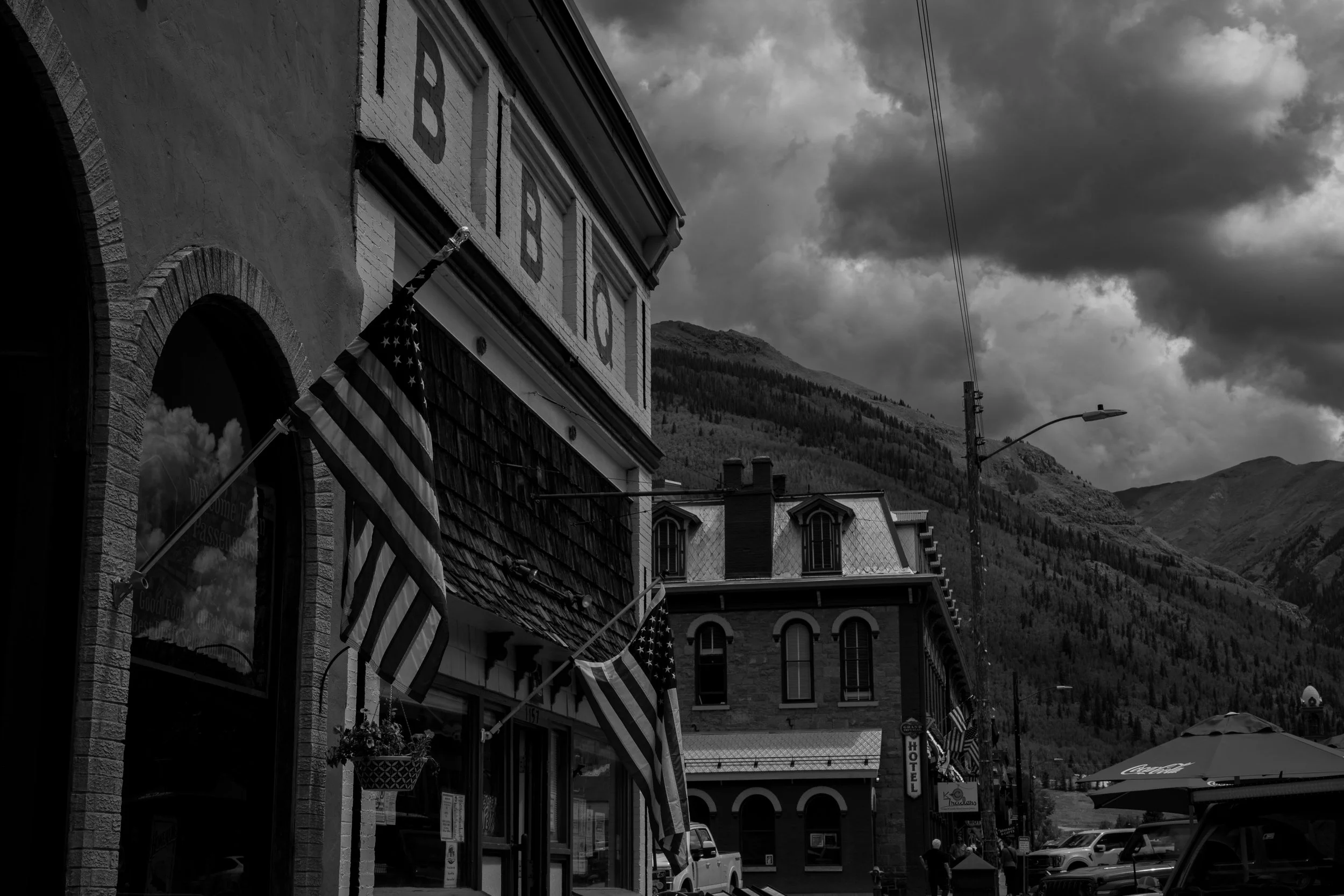 Street view of buildings with American flags, parked cars, and mountain backdrop under cloudy sky.