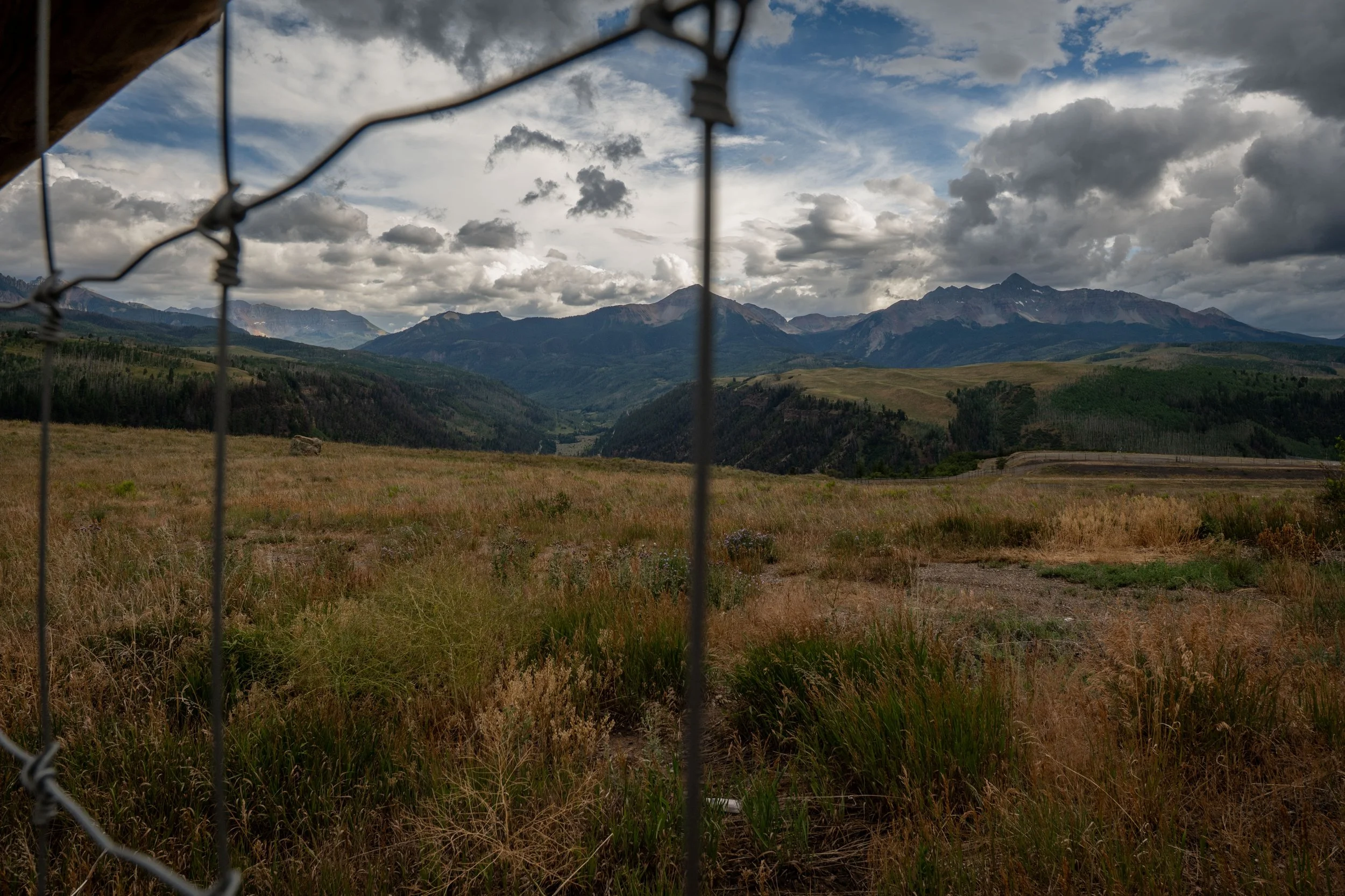 View of mountains and cloudy sky seen through a chain-link fence with some barbed wire at the top.