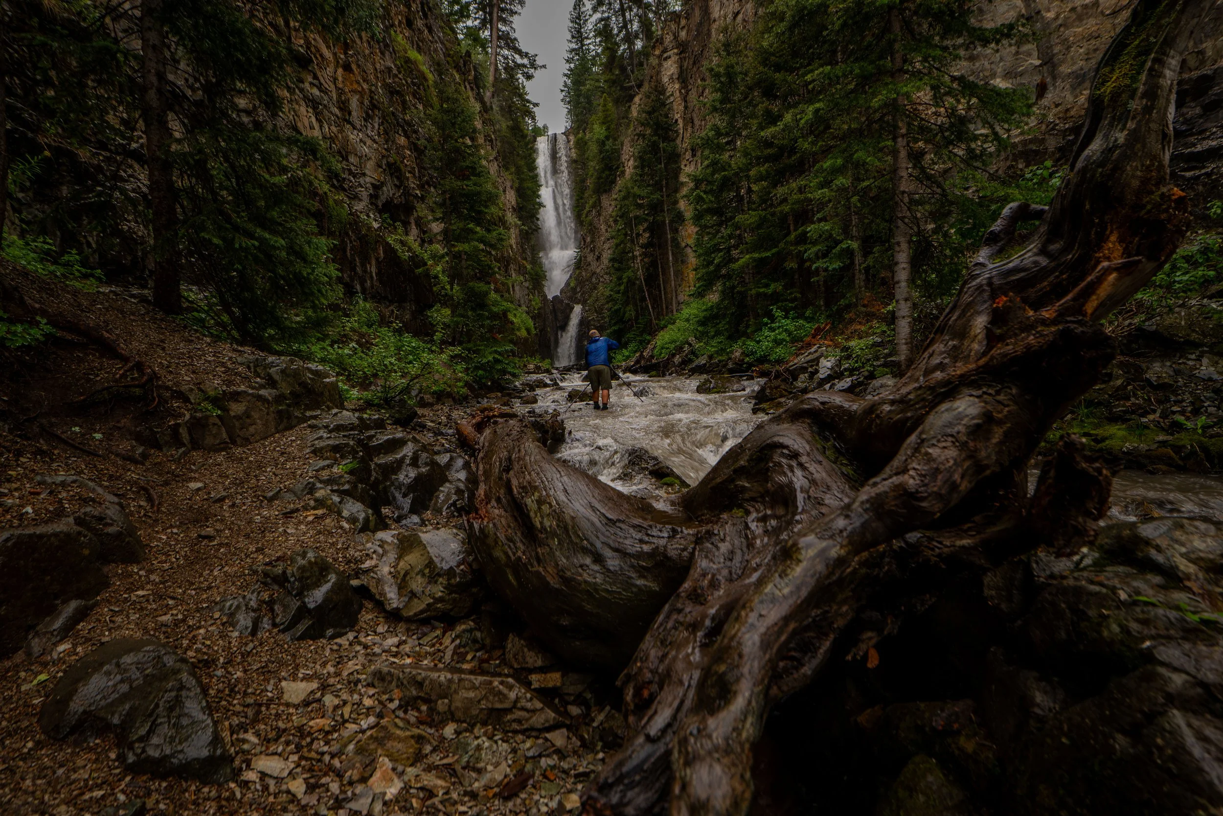 A person in a blue jacket with a hat, standing in a river with a waterfall in the background, surrounded by lush green trees and rocky terrain.