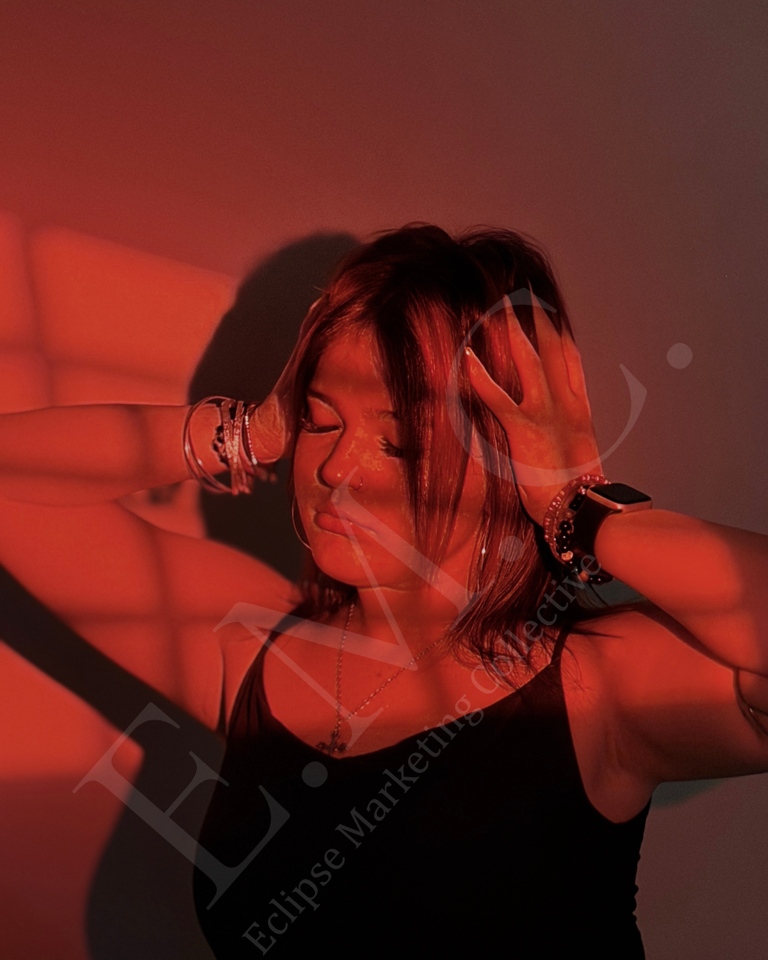 A woman with short dark hair, wearing a black tank top, jewelry, a smartwatch, and a nose ring, posing with her hands in her hair against a plain wall, illuminated by red and orange lighting.