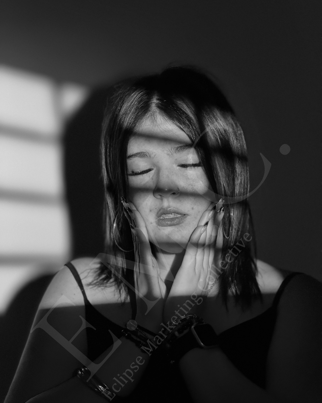 Black and white portrait of a young woman with closed eyes, gently touching her face with both hands, with shadows and light creating a dramatic effect.