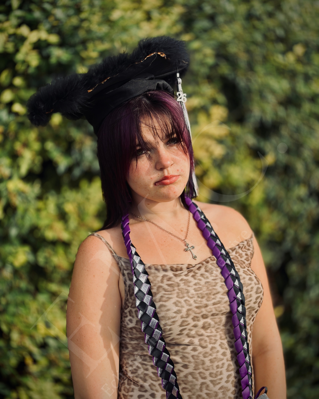 A woman with purple and black braided hair, wearing a leopard print dress and a large black hat with feathers, standing outdoors with green foliage in the background.