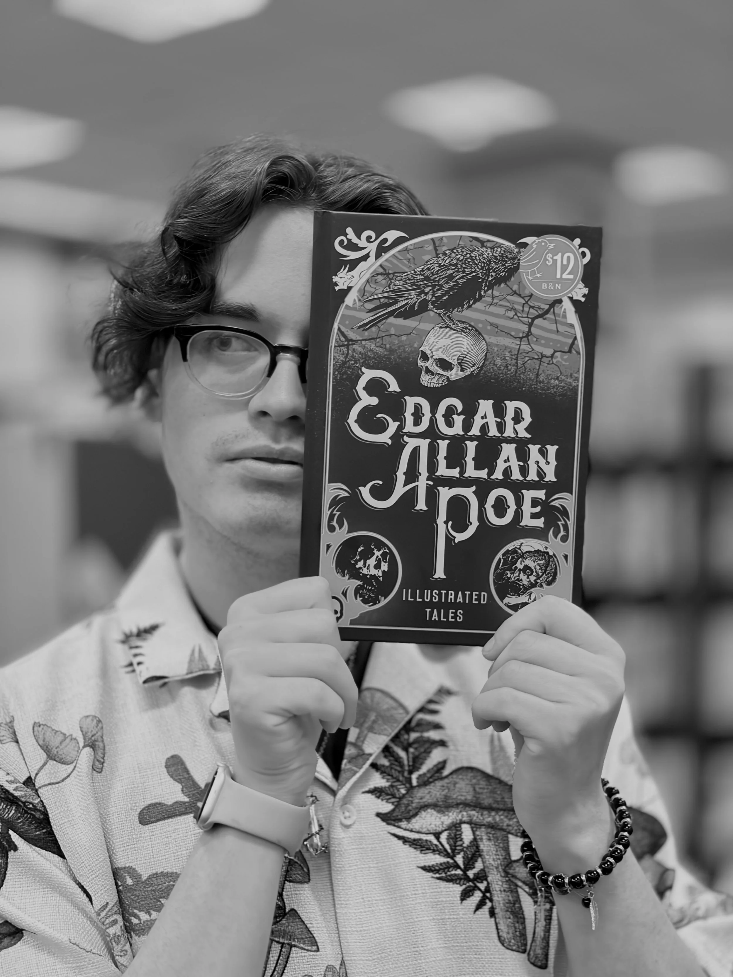 A person with glasses holding a book titled 'Edgar Allan Poe: Illustrated Tales' in front of their face, with only one eye visible. They are wearing a shirt with a floral pattern and a beaded bracelet.