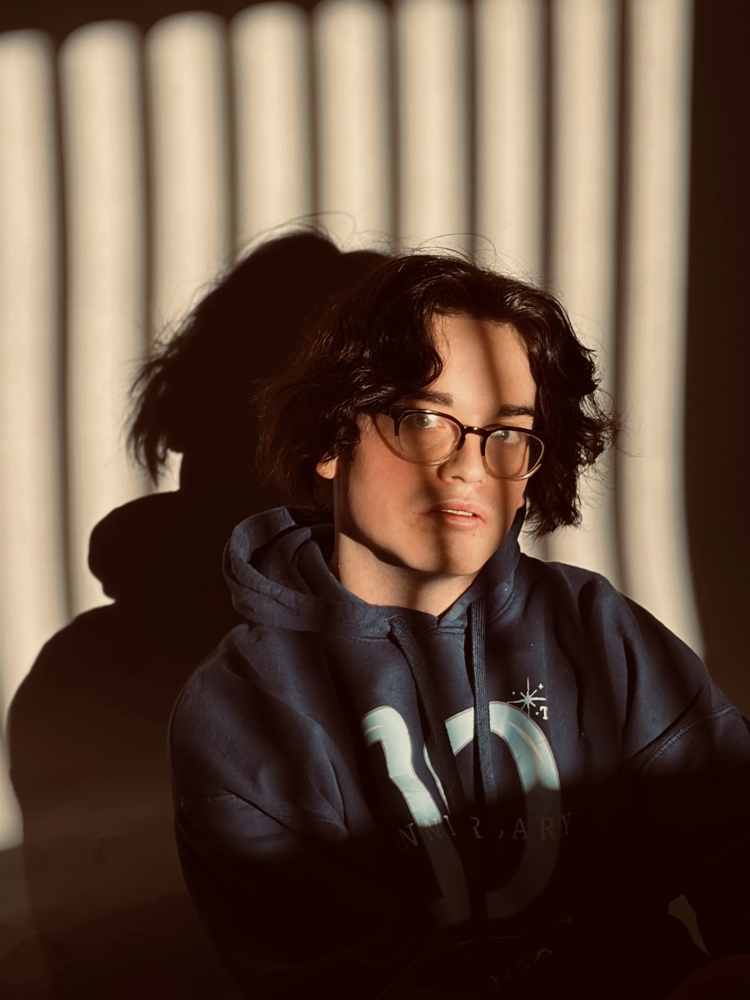 A person with dark, curly hair wearing glasses and a dark hoodie, looking slightly to the side, sitting in front of a background with vertical beige panels.