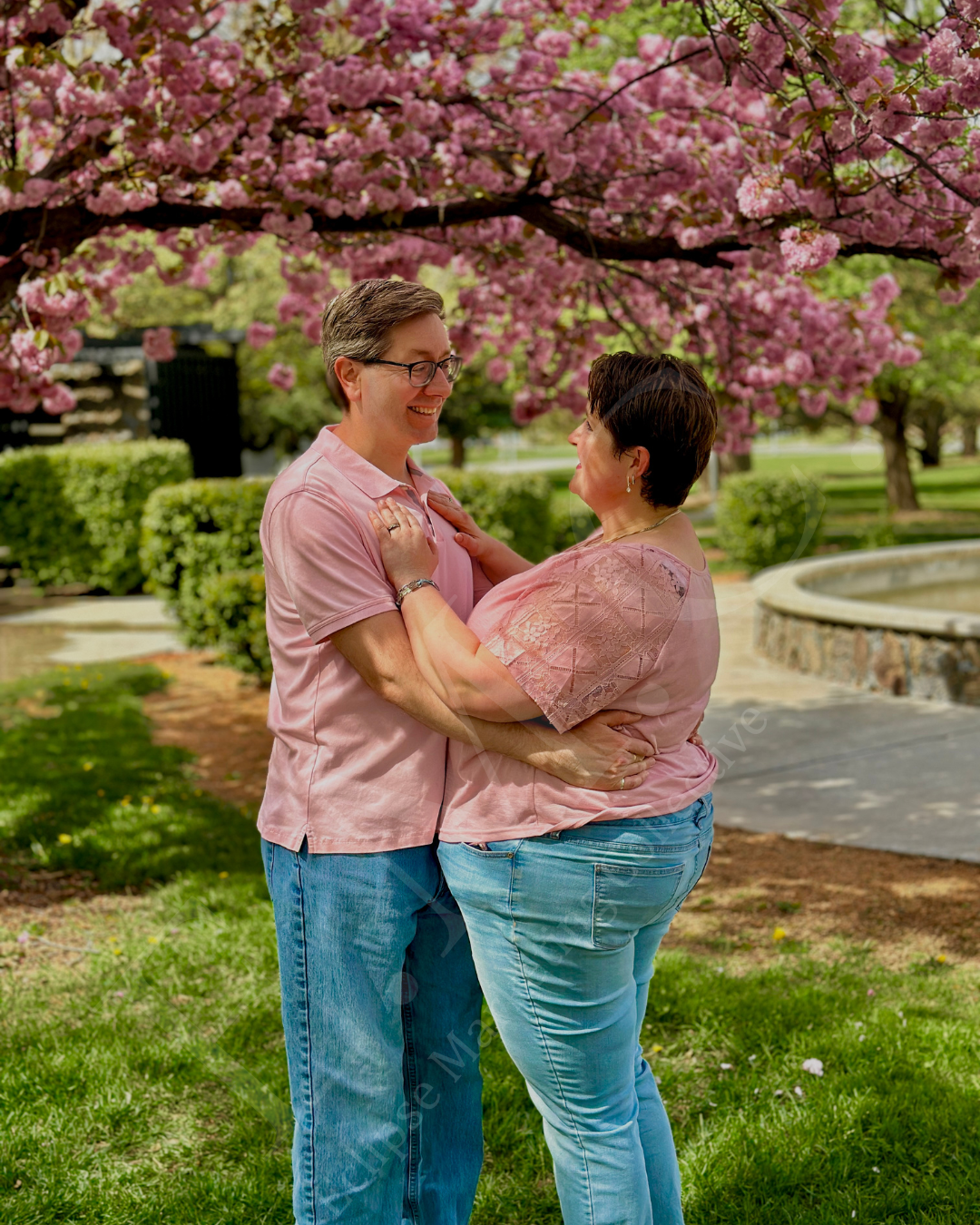 A couple stands close together under blooming pink cherry blossom trees in a park, smiling at each other.