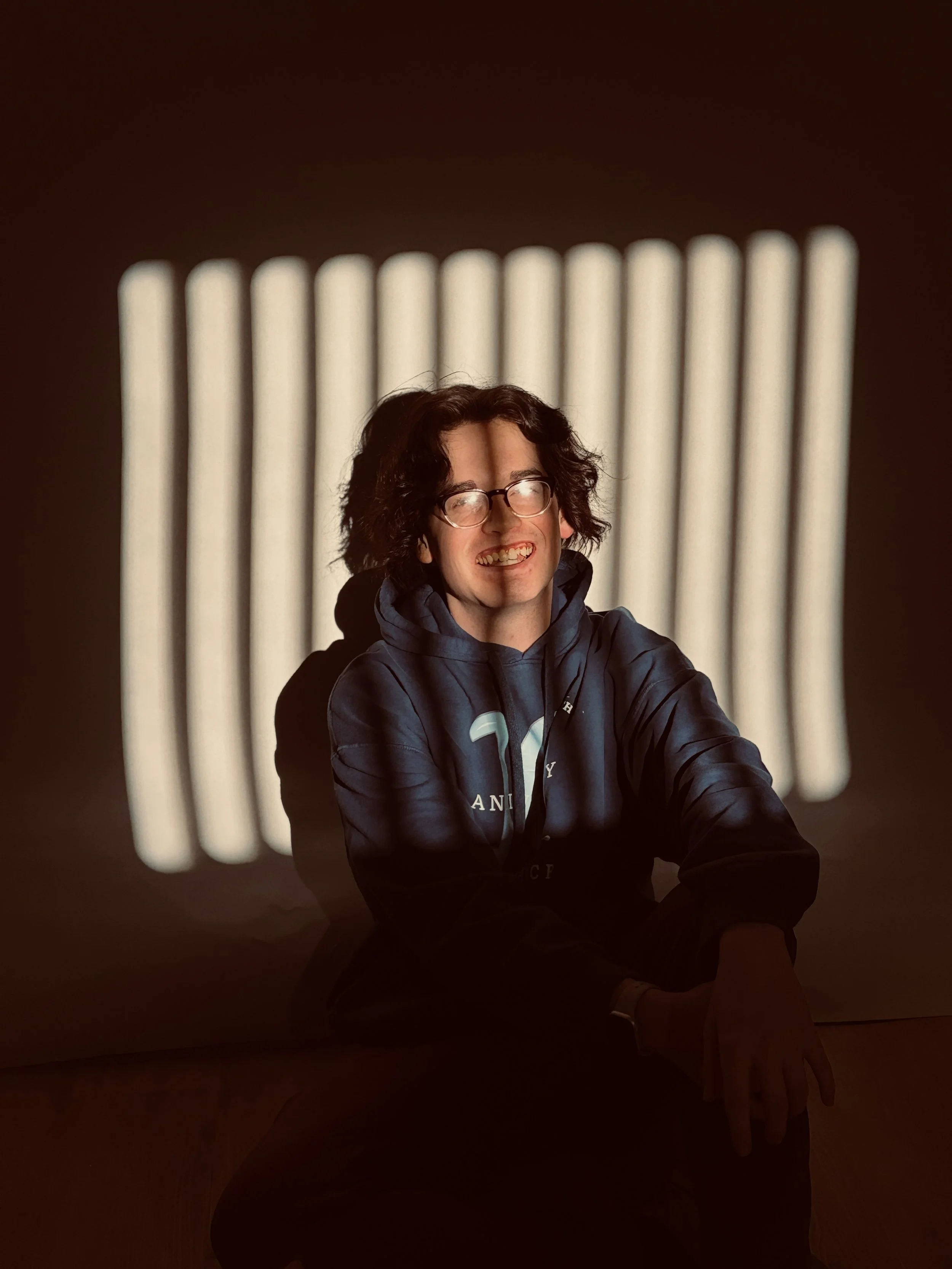 A person with curly hair and glasses smiling, sitting in front of a wall with vertical white light patterns.