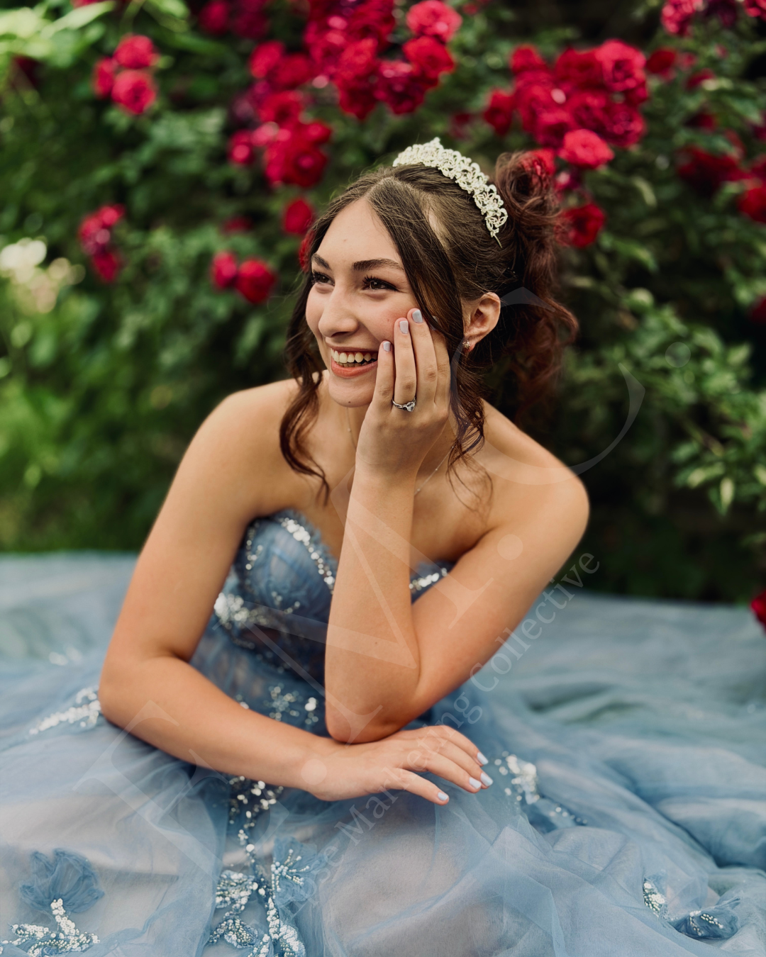 A young woman in a strapless blue dress with sequins, sitting outdoors on a stone surface, smiling and touching her cheek, with pink flowers and green foliage in the background.
