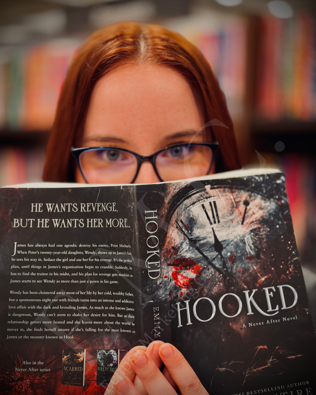 A woman with red hair and glasses is reading a dark-themed book titled "Hooked" in a library with blurred bookshelves in the background.