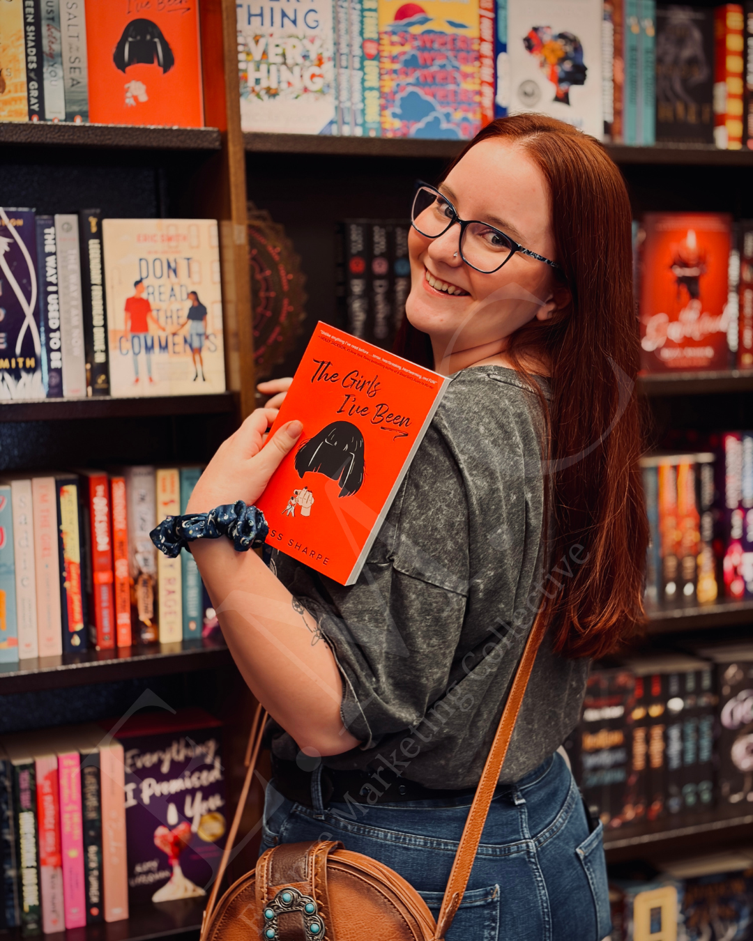 A young woman with red hair, glasses, and a nose ring at a bookstore holding a book titled 'The Girls I've Been' close to her chest and smiling at the camera.