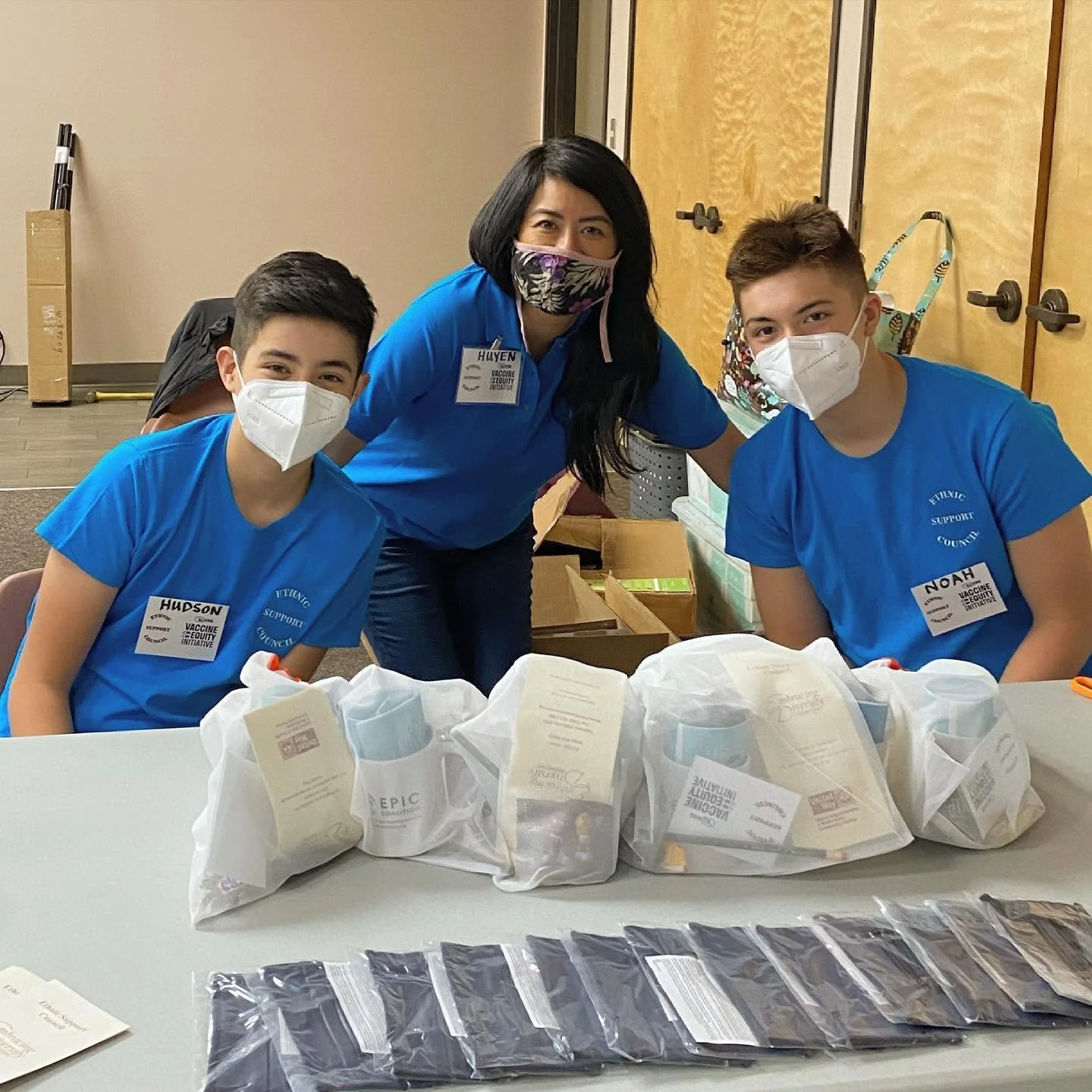 Three masked volunteers in blue shirts pose behind packaged mugs and supplies at a vaccine equity initiative event.