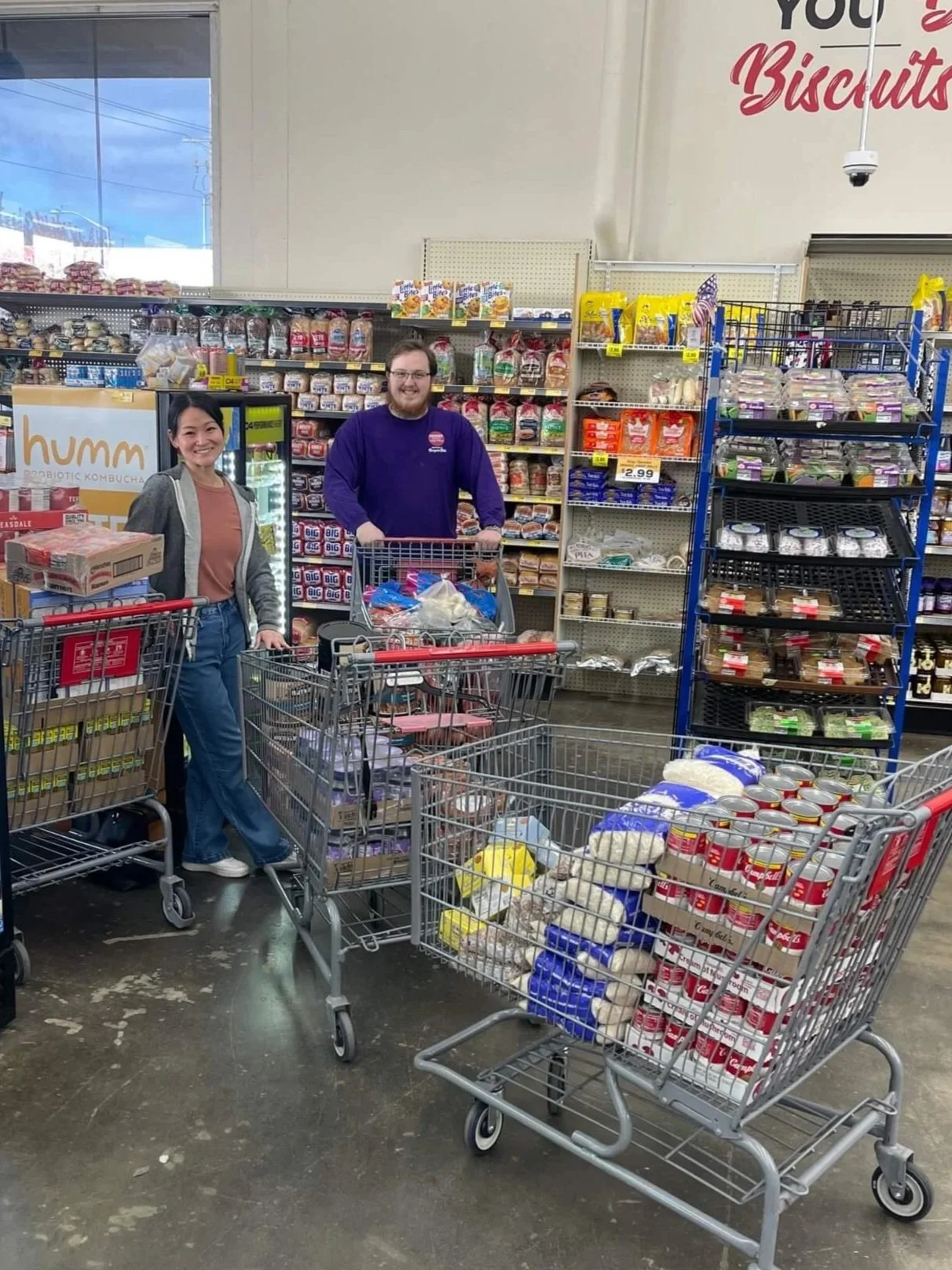 Two shoppers with full grocery carts inside a supermarket, surrounded by shelves of bread, meat, dairy products, and canned goods.