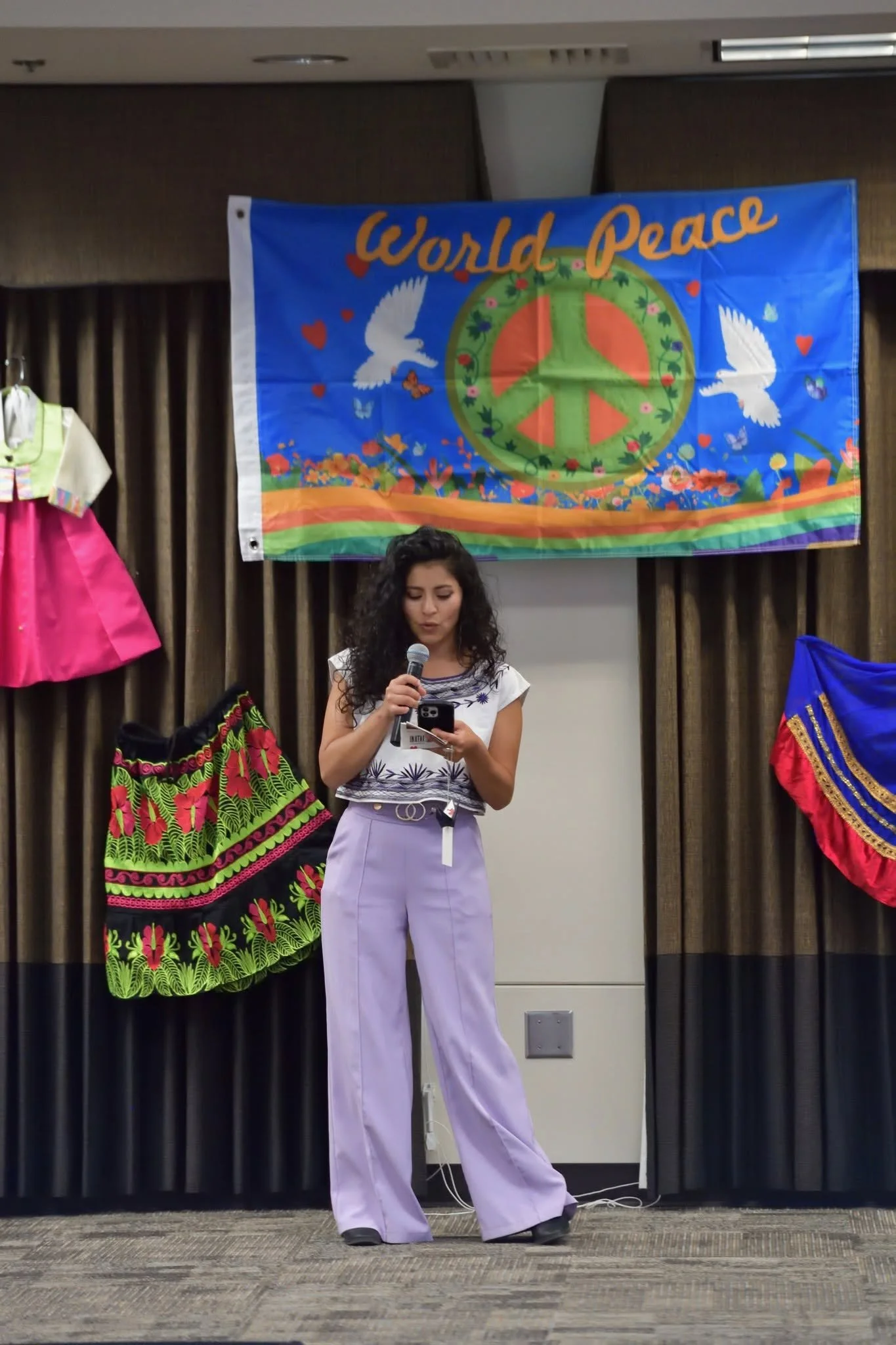 Woman speaking into a microphone beneath a colorful World Peace banner at a cultural event