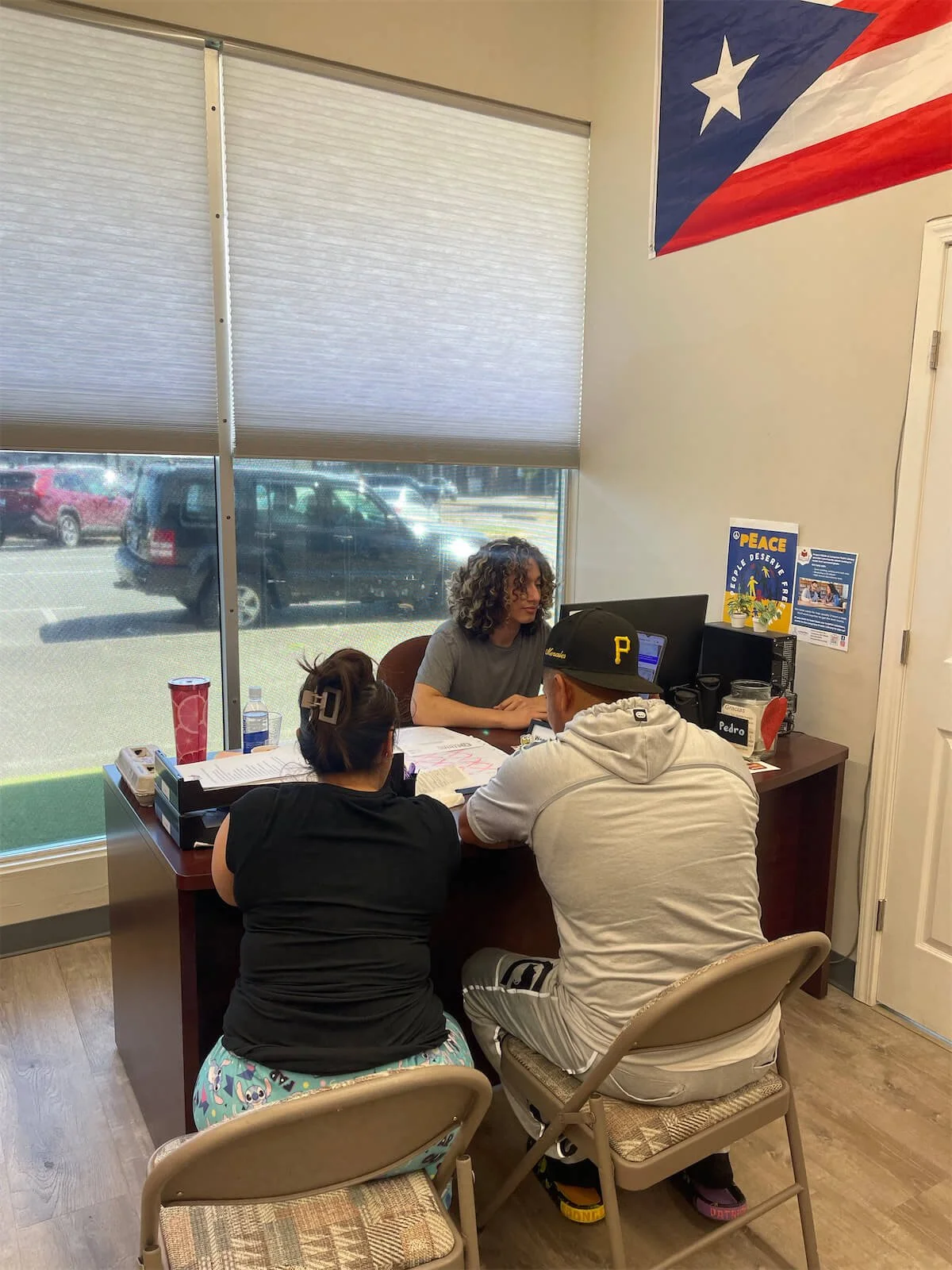 Three people meet at a desk in an office beneath a Puerto Rican flag, with paperwork and a computer.